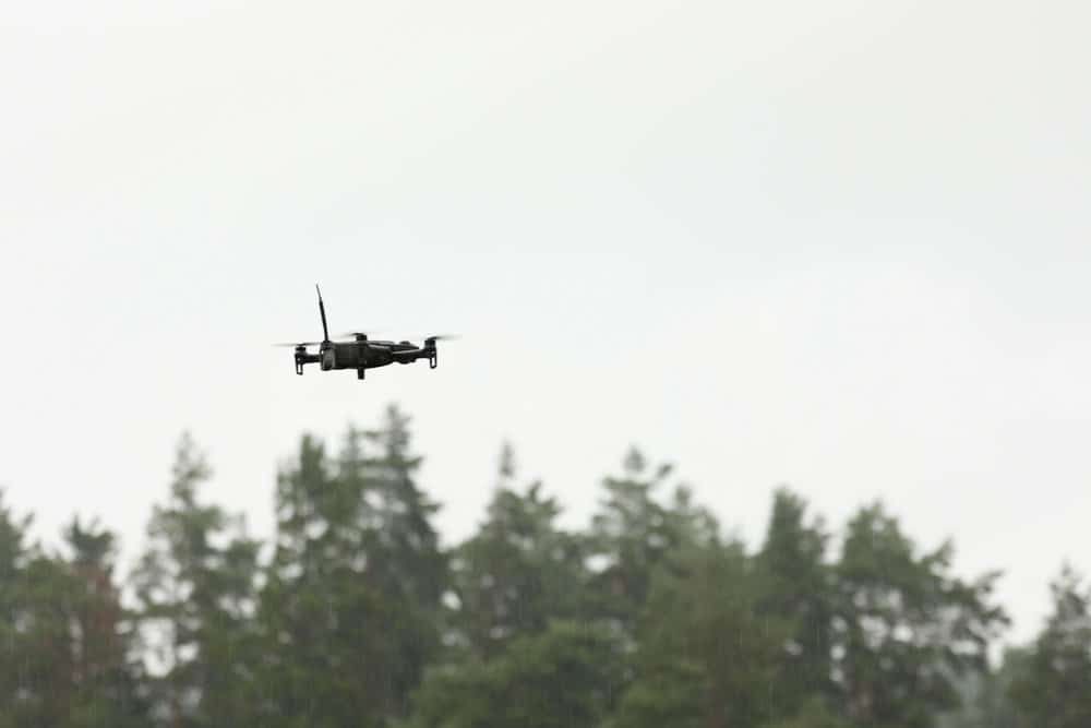 A small quadcopter flies above a conifer treeline under heavy overcast.