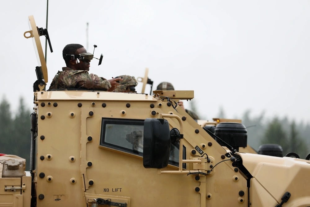 A soldier wearing FPV goggles sits in the turret hatch of a tan armored truck.