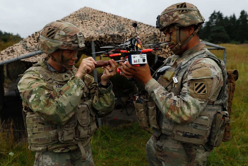 Two soldiers examine a red racing quadcopter beneath a camouflage net in a field.