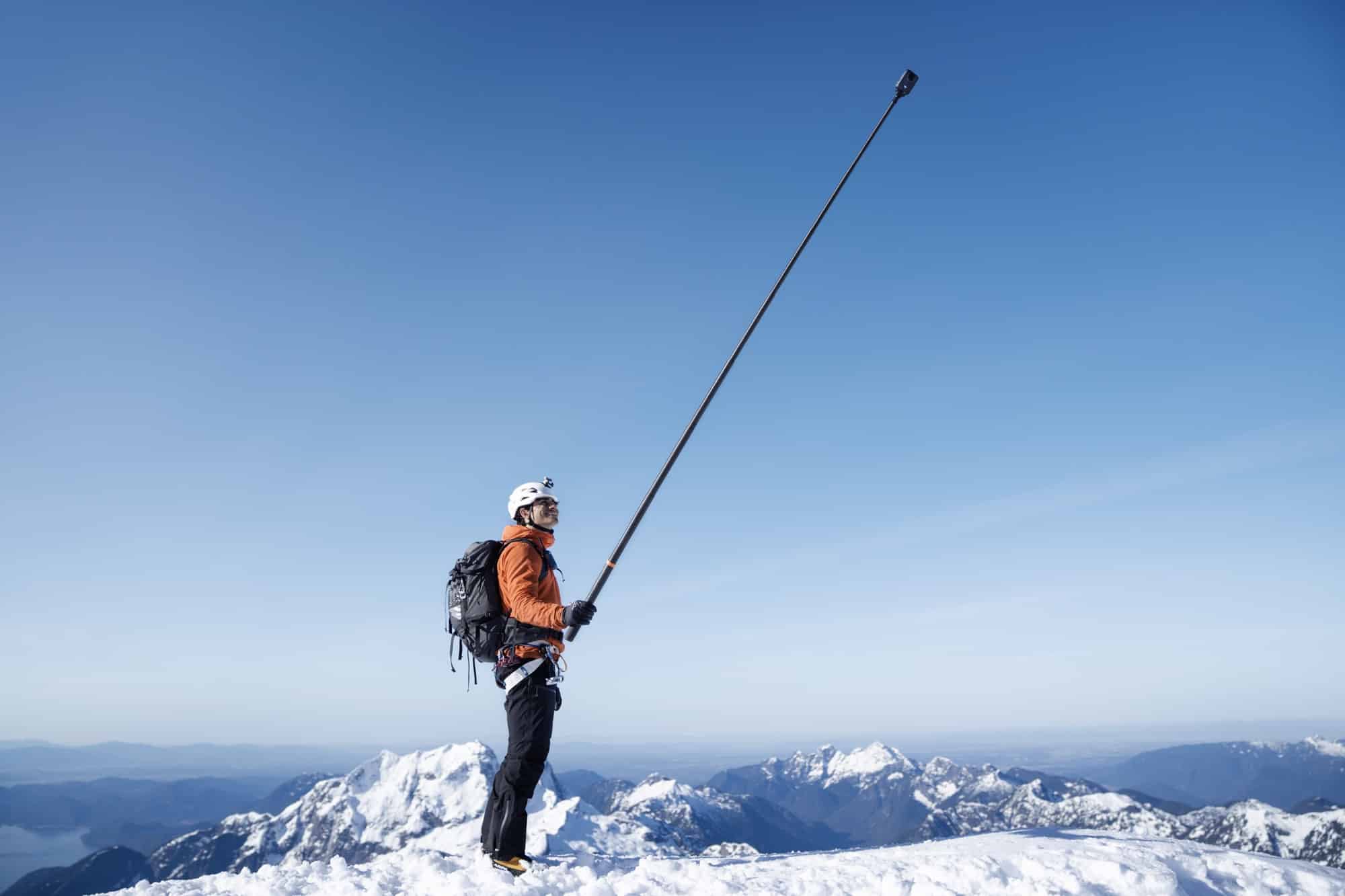 A mountaineer on a snowy summit extends a long carbon fiber selfie stick skyward.