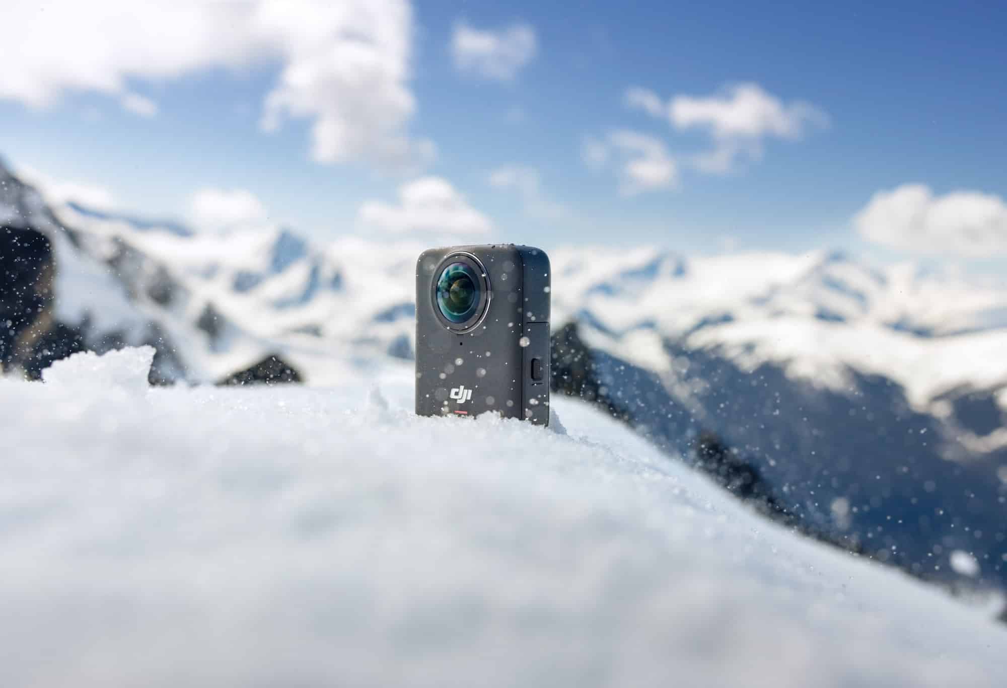 Close-up of the Osmo 360 half-buried in snow with alpine peaks blurred behind.