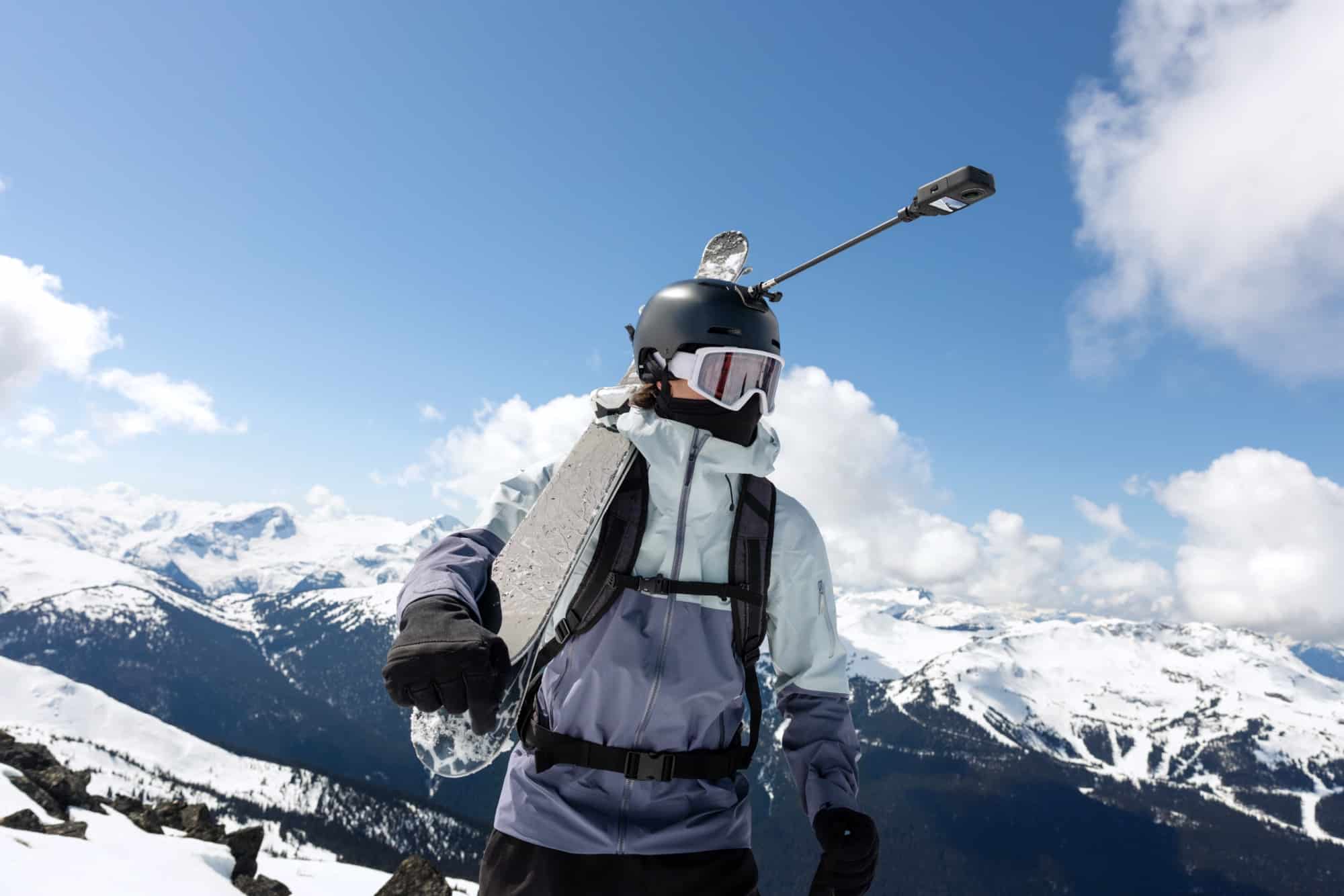 A skier with a helmet-mounted extension arm looks back across glaciated peaks.