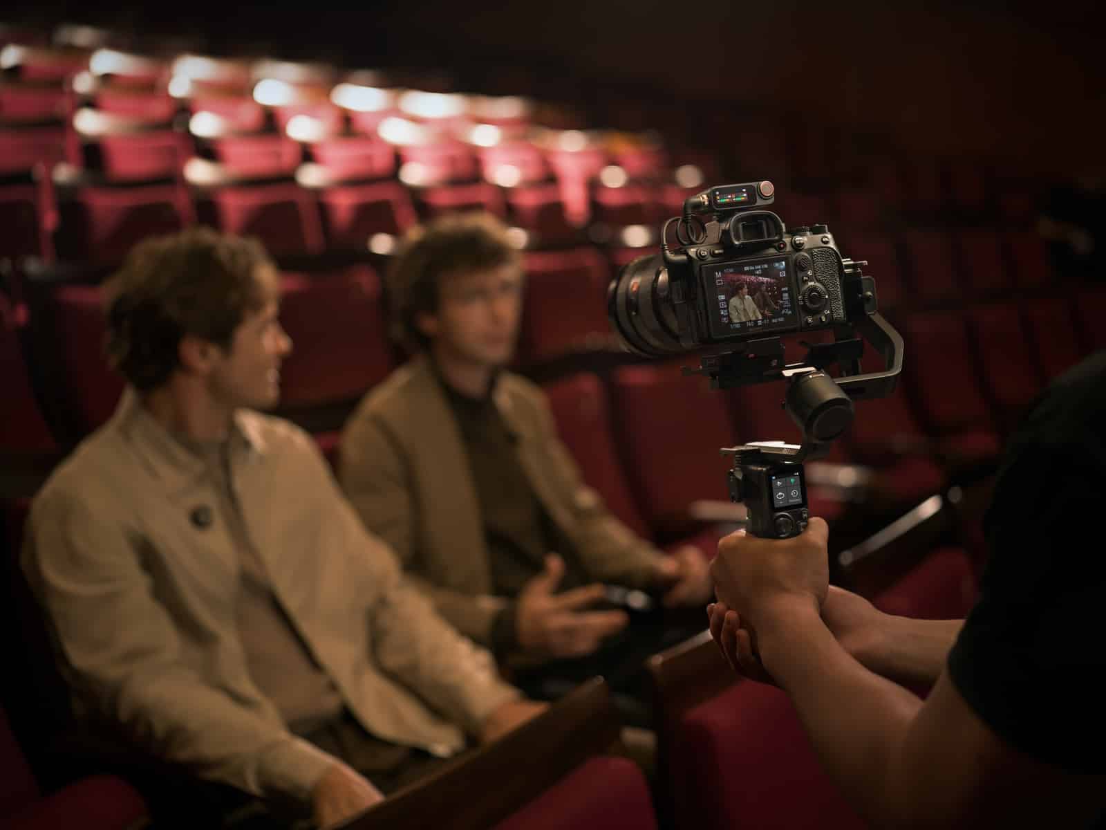 Two men converse in red theater seats while a gimbal rig records them.