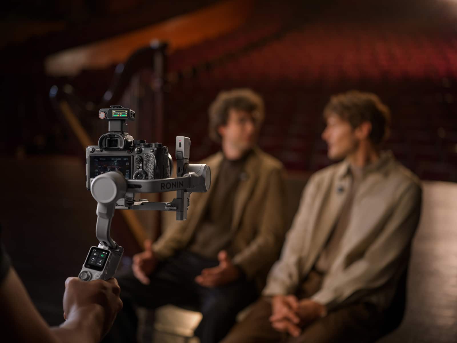 Gimbal-mounted camera in the foreground frames two men in beige coats.