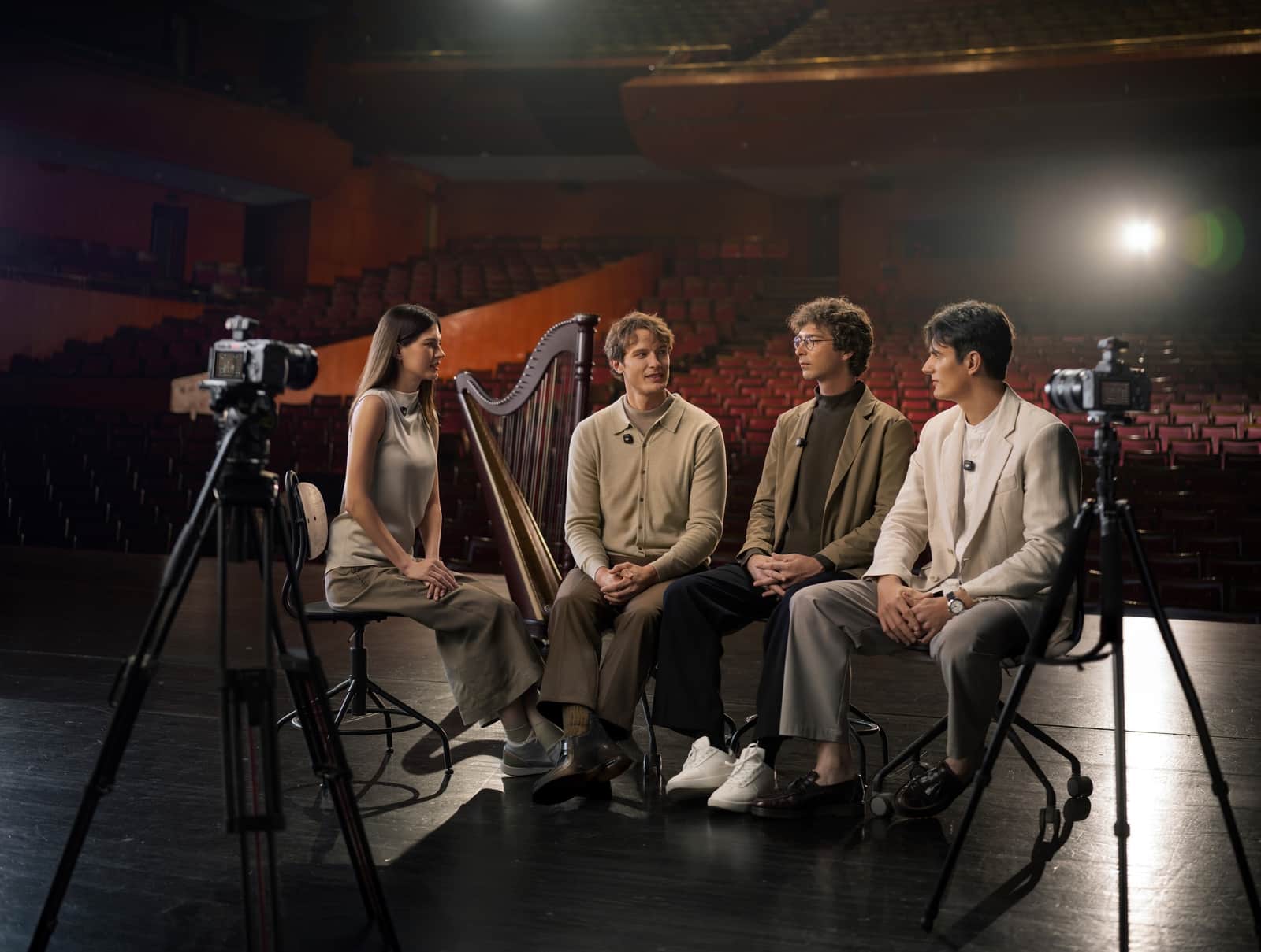 Four performers with a harp sit on stage between two camera tripods.