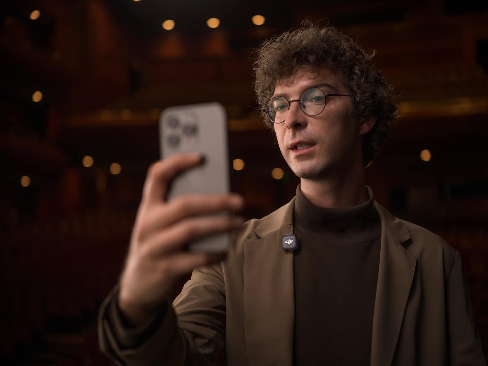Curly-haired man raises a phone toward himself in a warmly lit hall.
