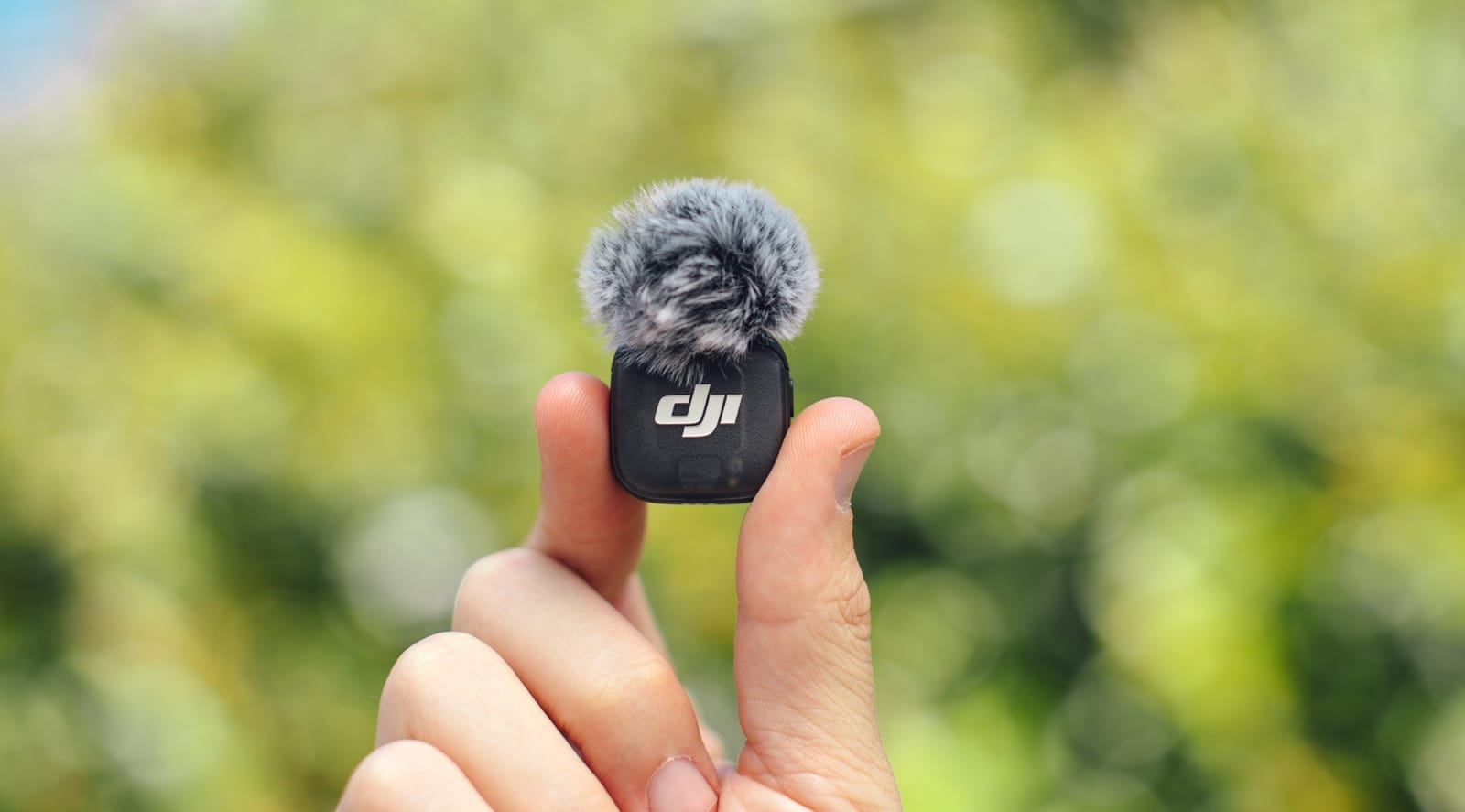 Fingers pinch a transmitter wearing a gray fur windscreen against blurred greenery.