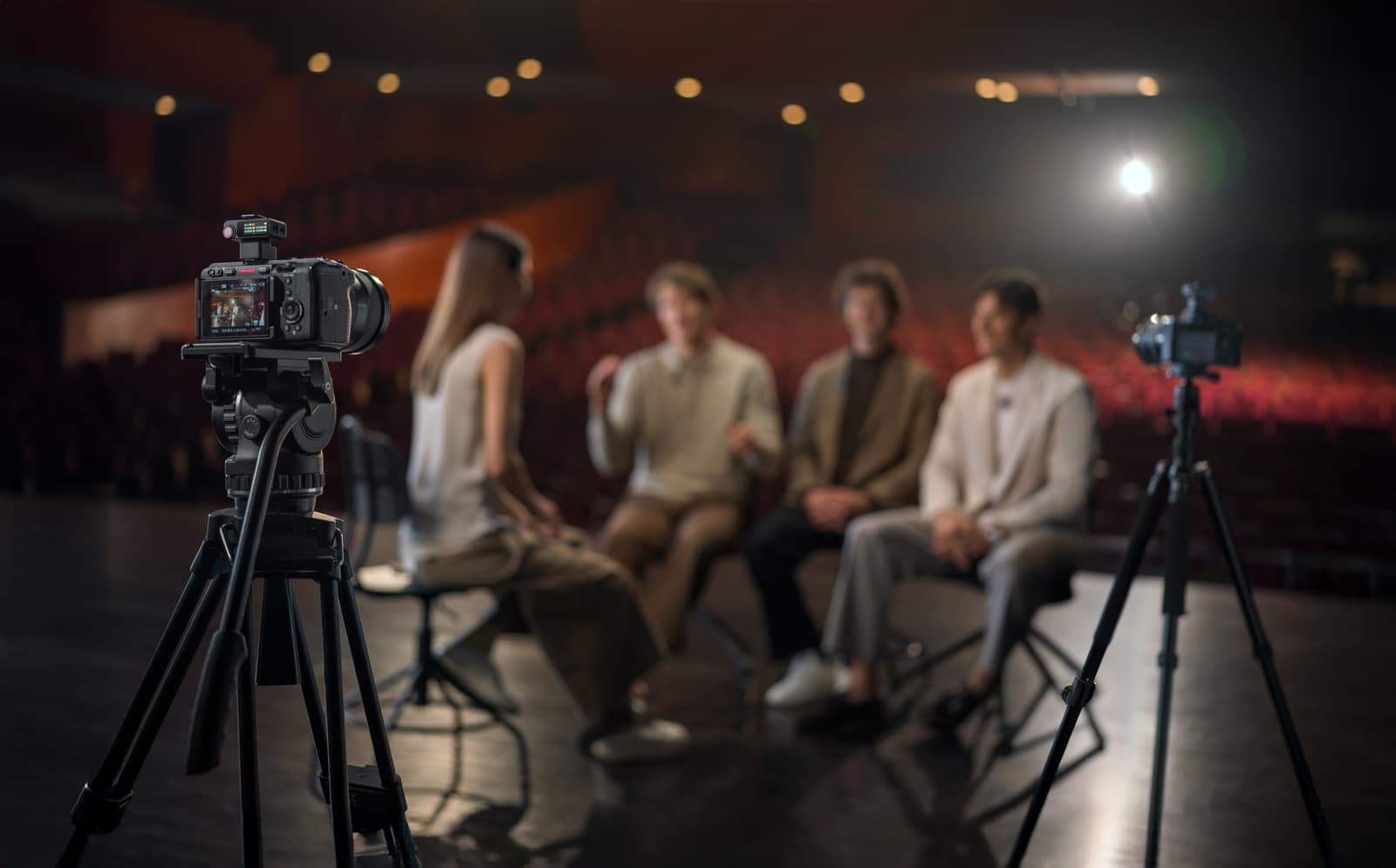 Two tripod cameras frame four people seated together on a theater stage.