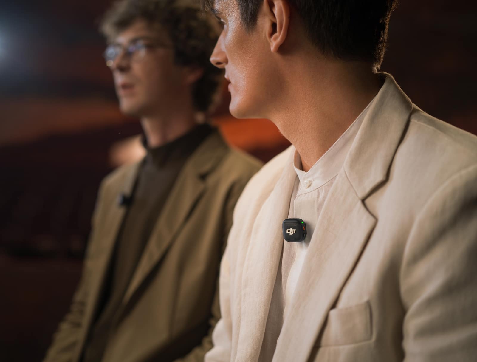 Two men seated in a dim theater talk, one wearing a transmitter.