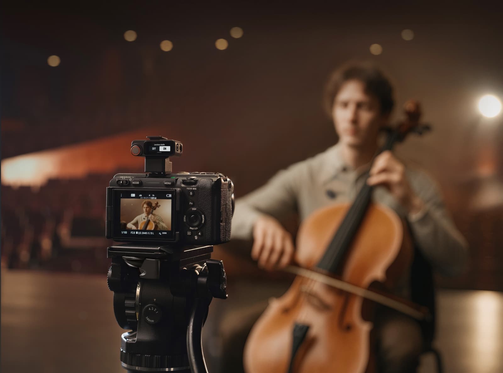 Tripod-mounted camera records a cellist performing in a warmly lit hall.