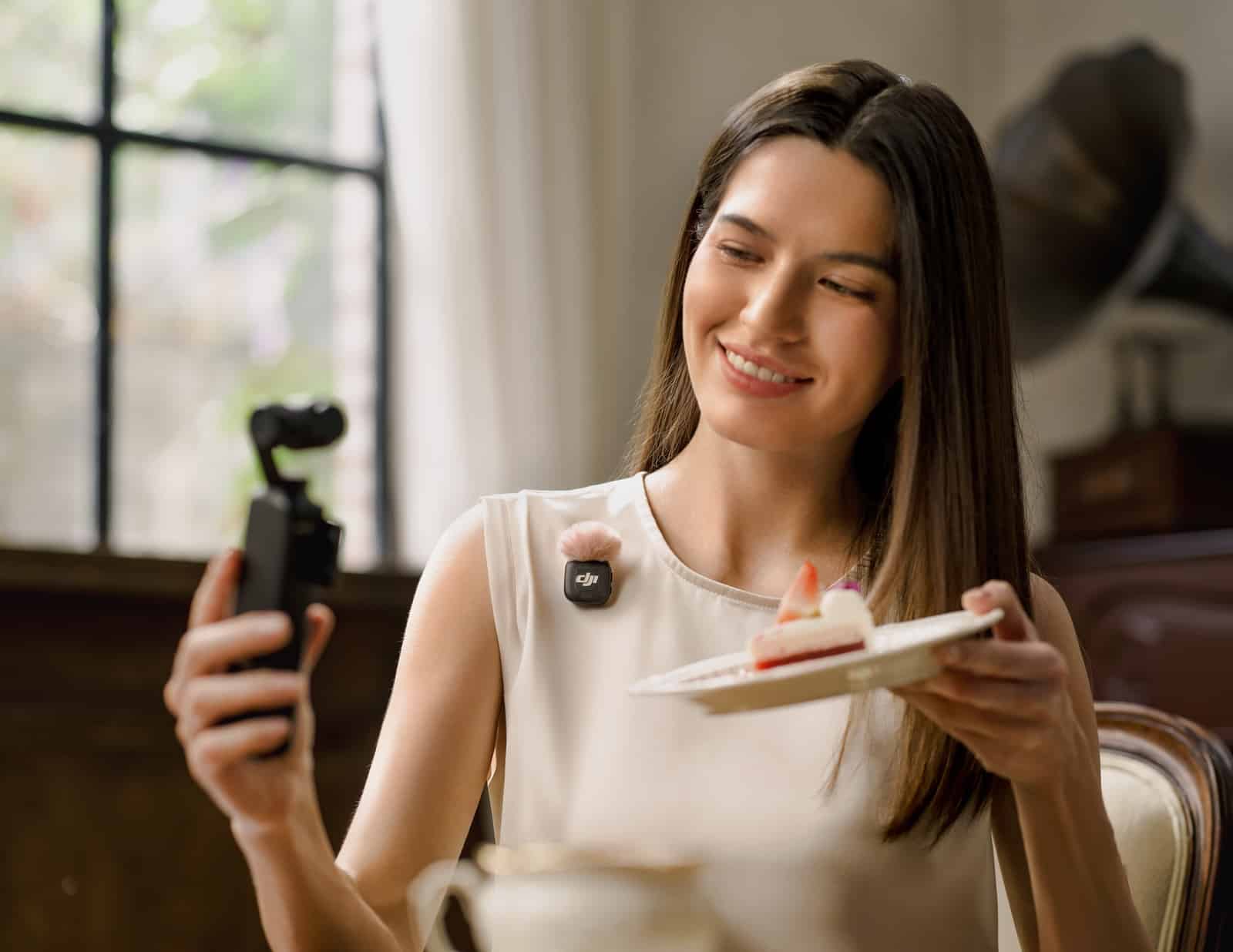 Woman holds a pocket camera and a plate of cake indoors.