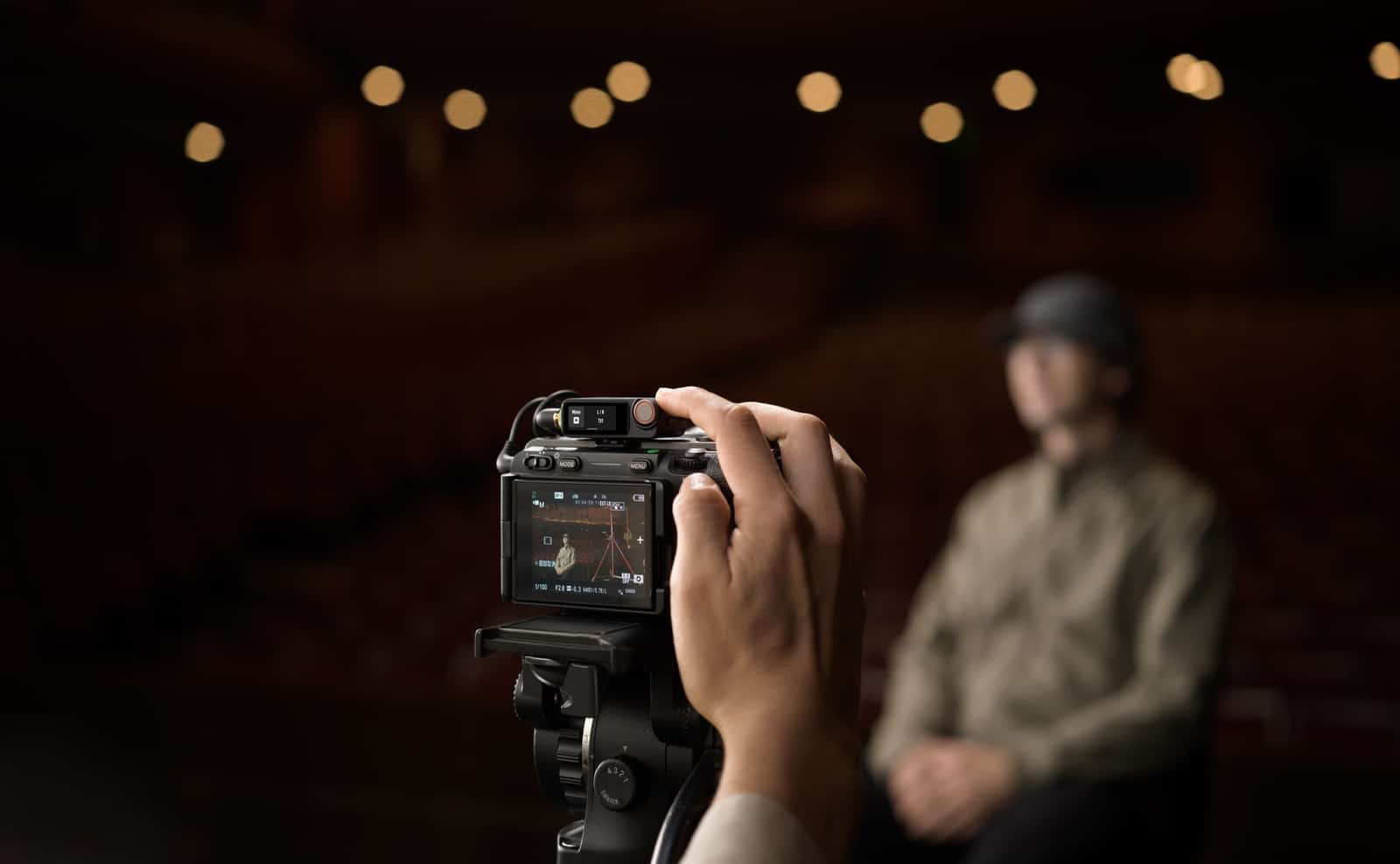 Operator adjusts a tripod camera aimed at a seated man in a theater.