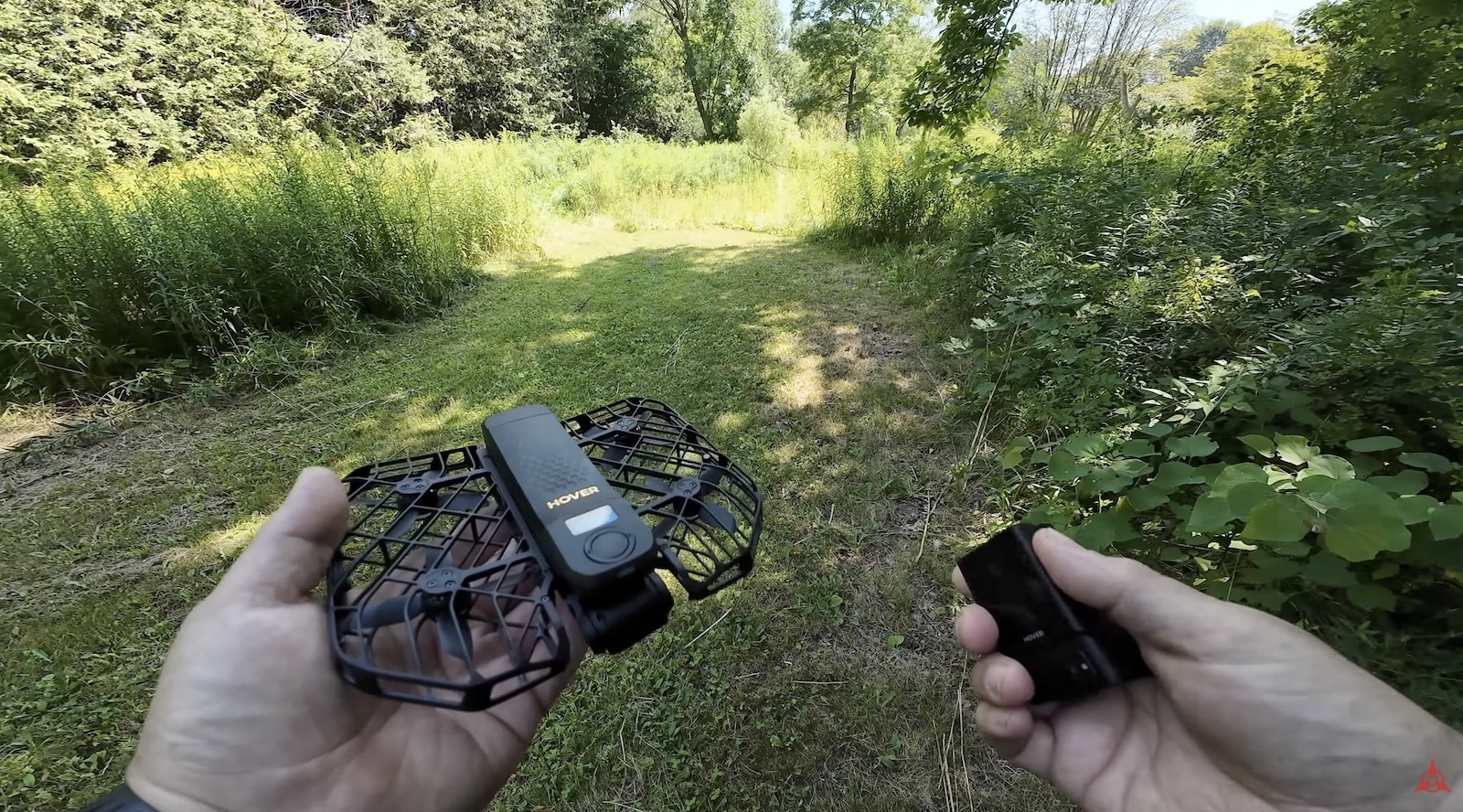 First-person view of hands holding a caged Hover Air X1 above a grassy trail.