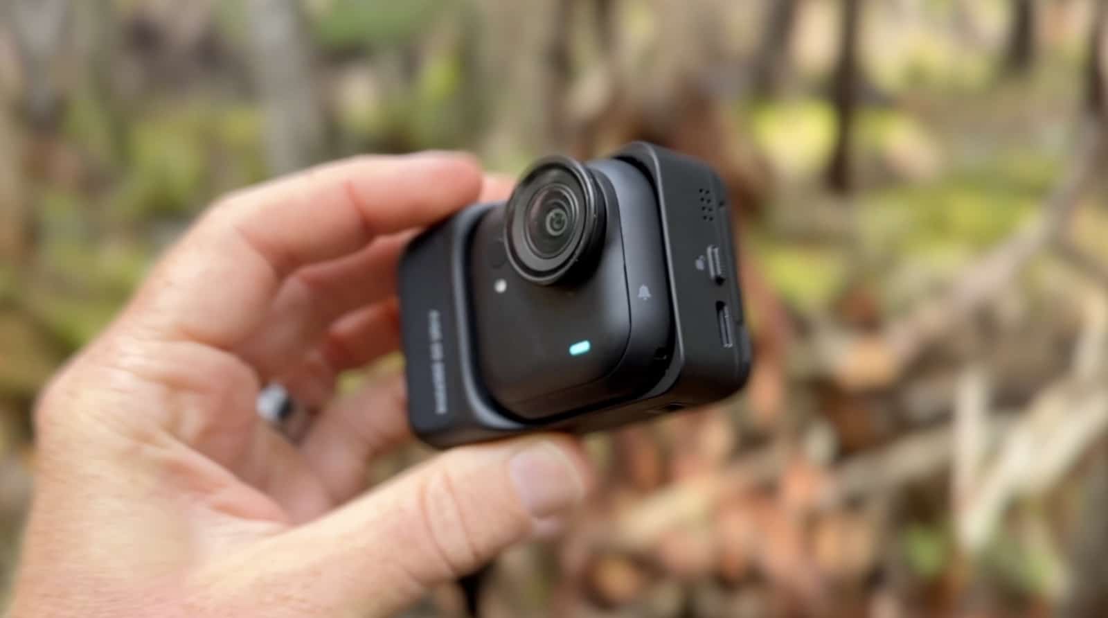 Hand holds the tiny Insta360 GO Ultra camera against blurred woodland.