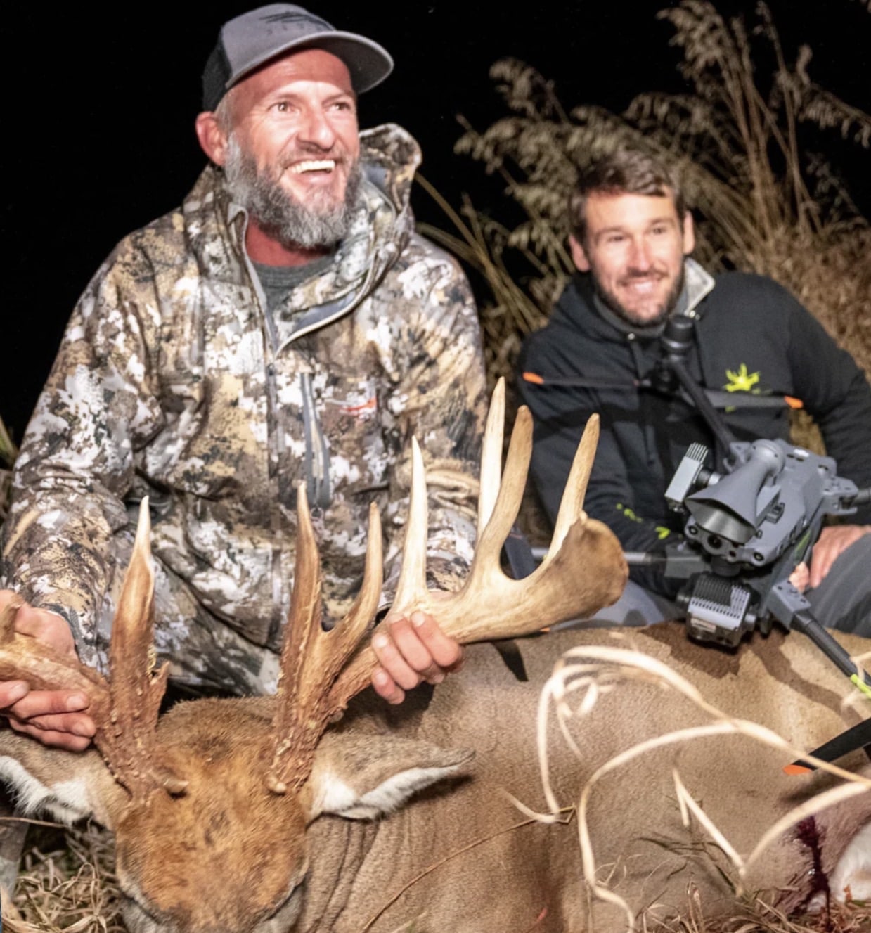 Two hunters kneel behind a large whitetail buck, one cradling a thermal drone.