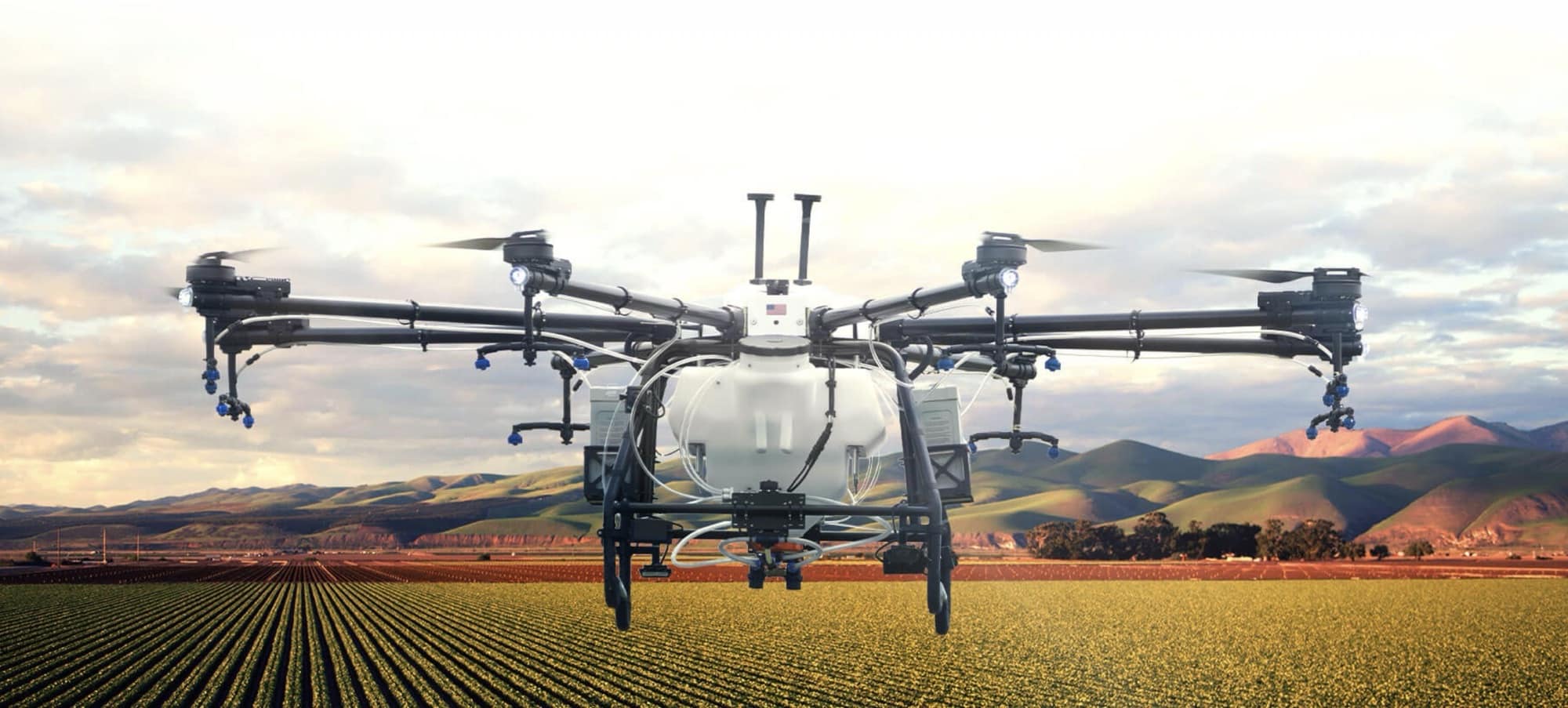 Spray drone hovering above crop rows at sunrise with mountains behind.