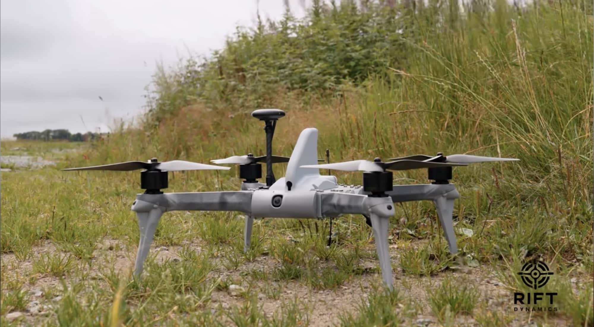 The Rift Dynamics Wasp quadcopter sits in tall grass with its antenna raised.