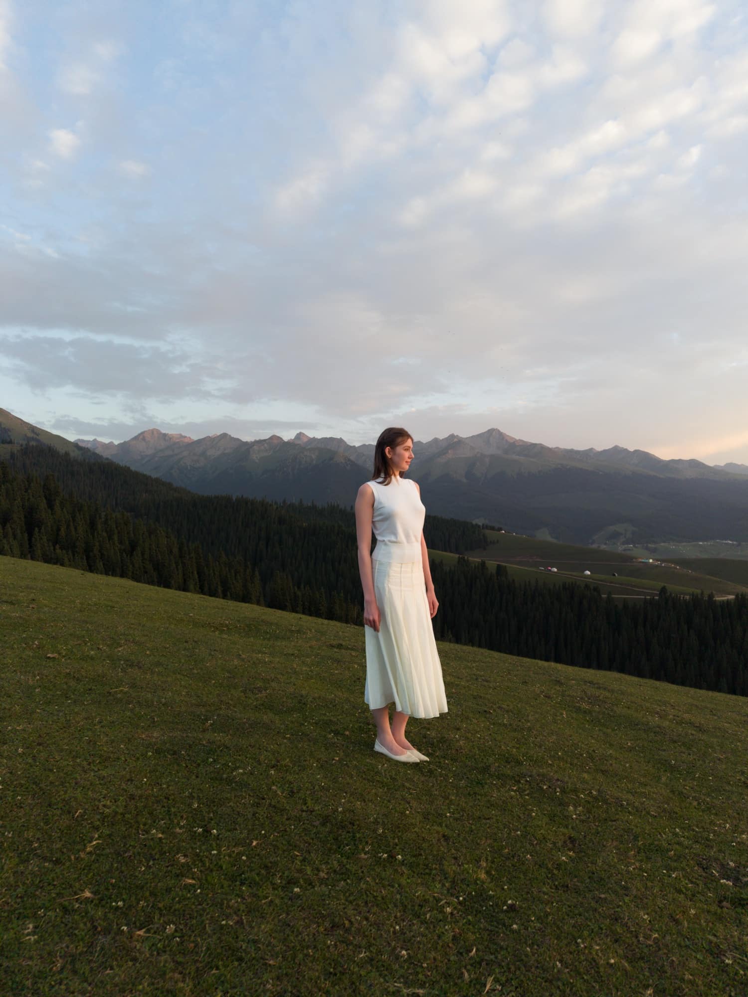 Wide framing of a woman in a long white dress on a dusk hillside.