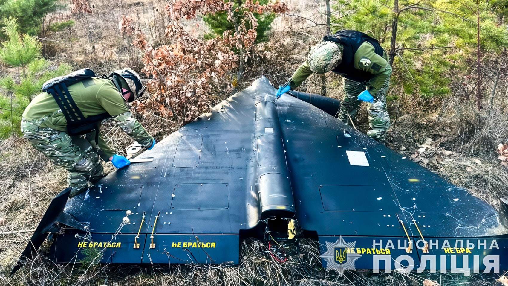 Ukrainian personnel inspecting a downed dark delta-wing drone in a forest clearing.