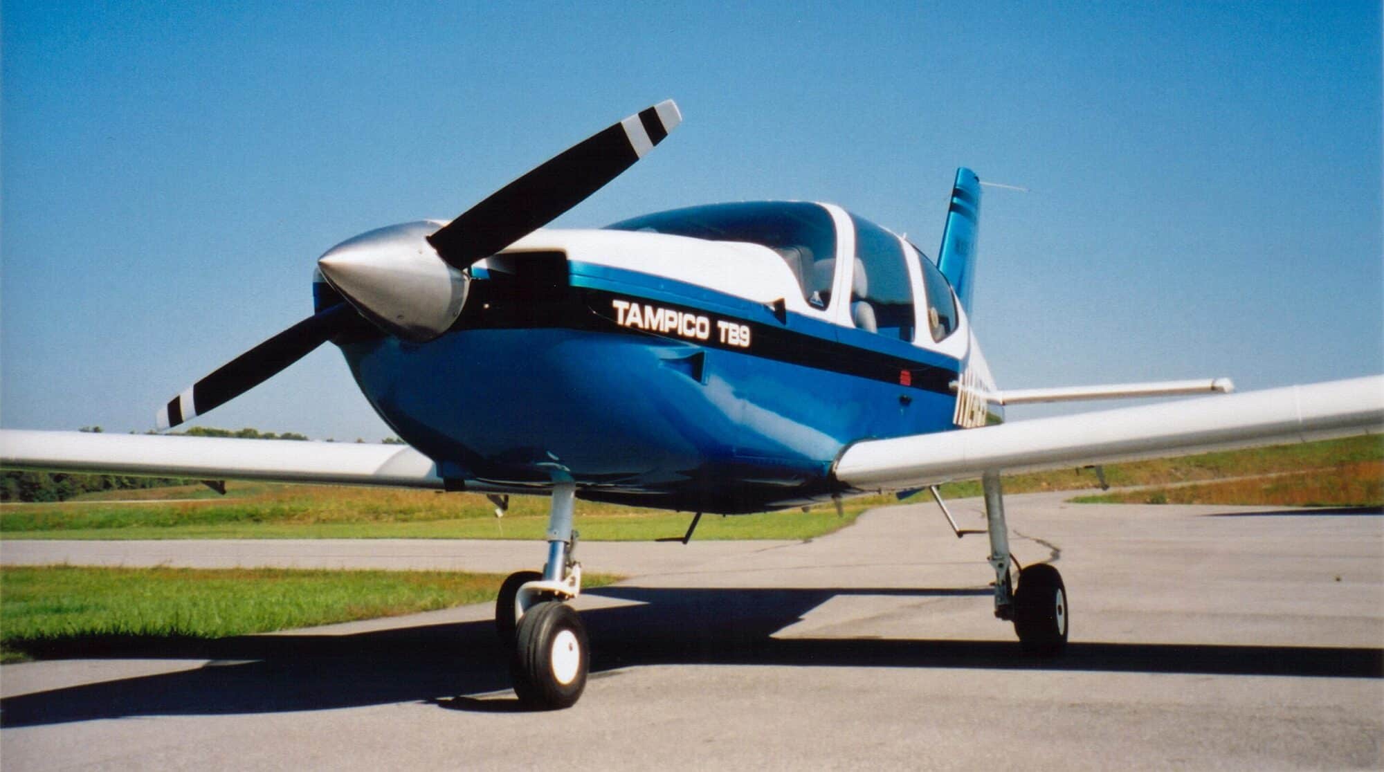 Blue and white Socata Tampico training aircraft parked beside a runway.