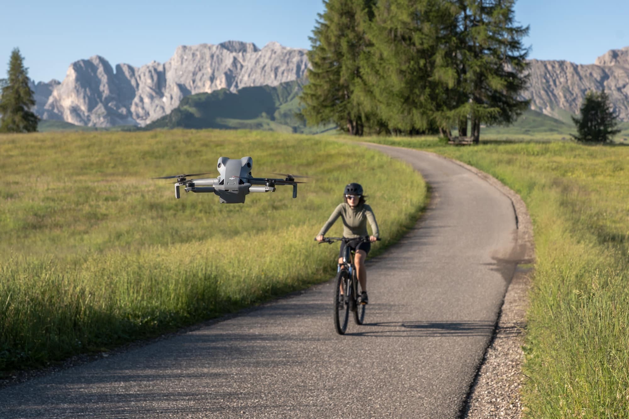 The drone flies alongside a cyclist on a paved path below jagged Dolomite peaks.