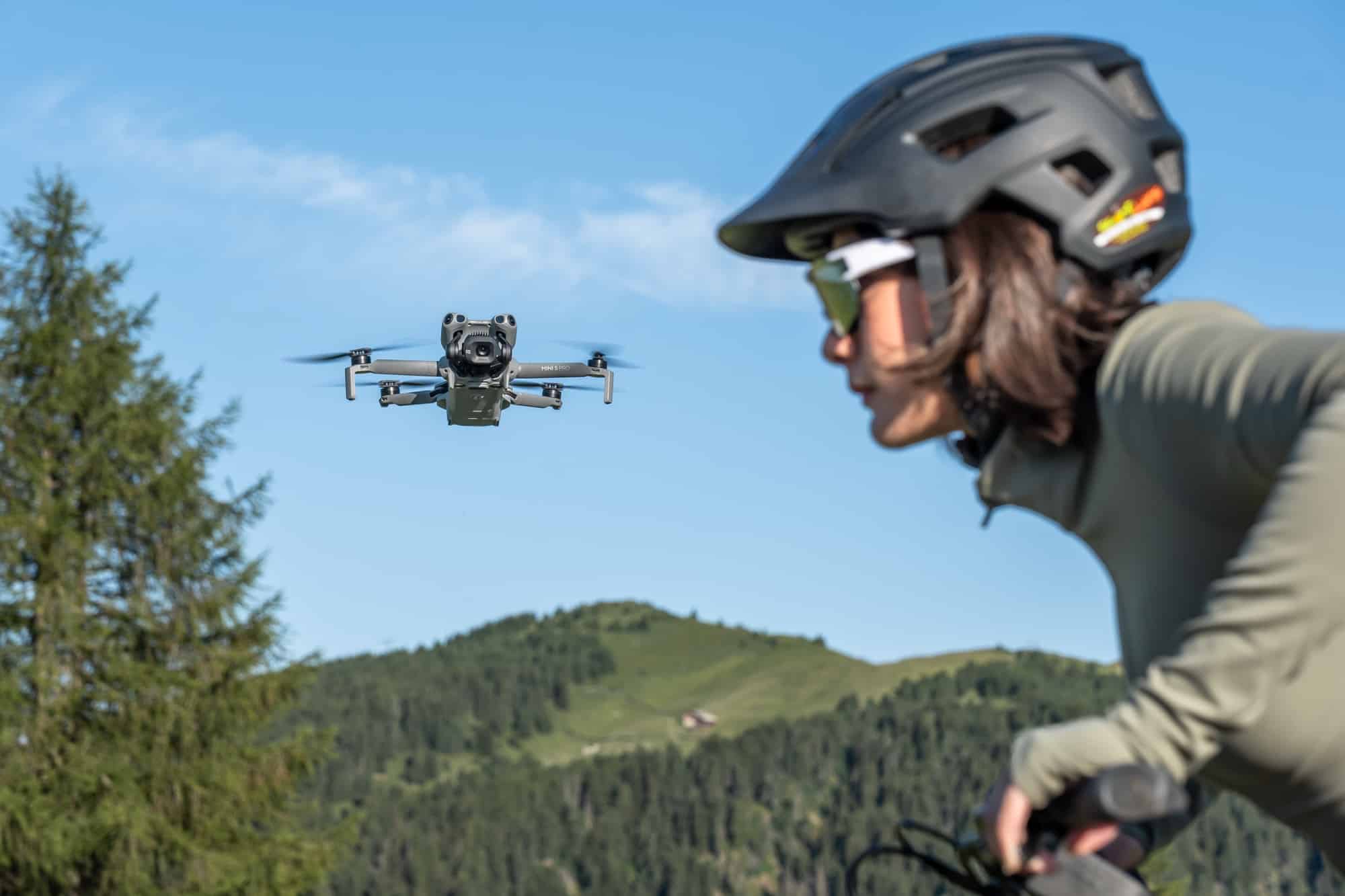 The drone flies close beside a helmeted rider with mountain slopes filling the background.