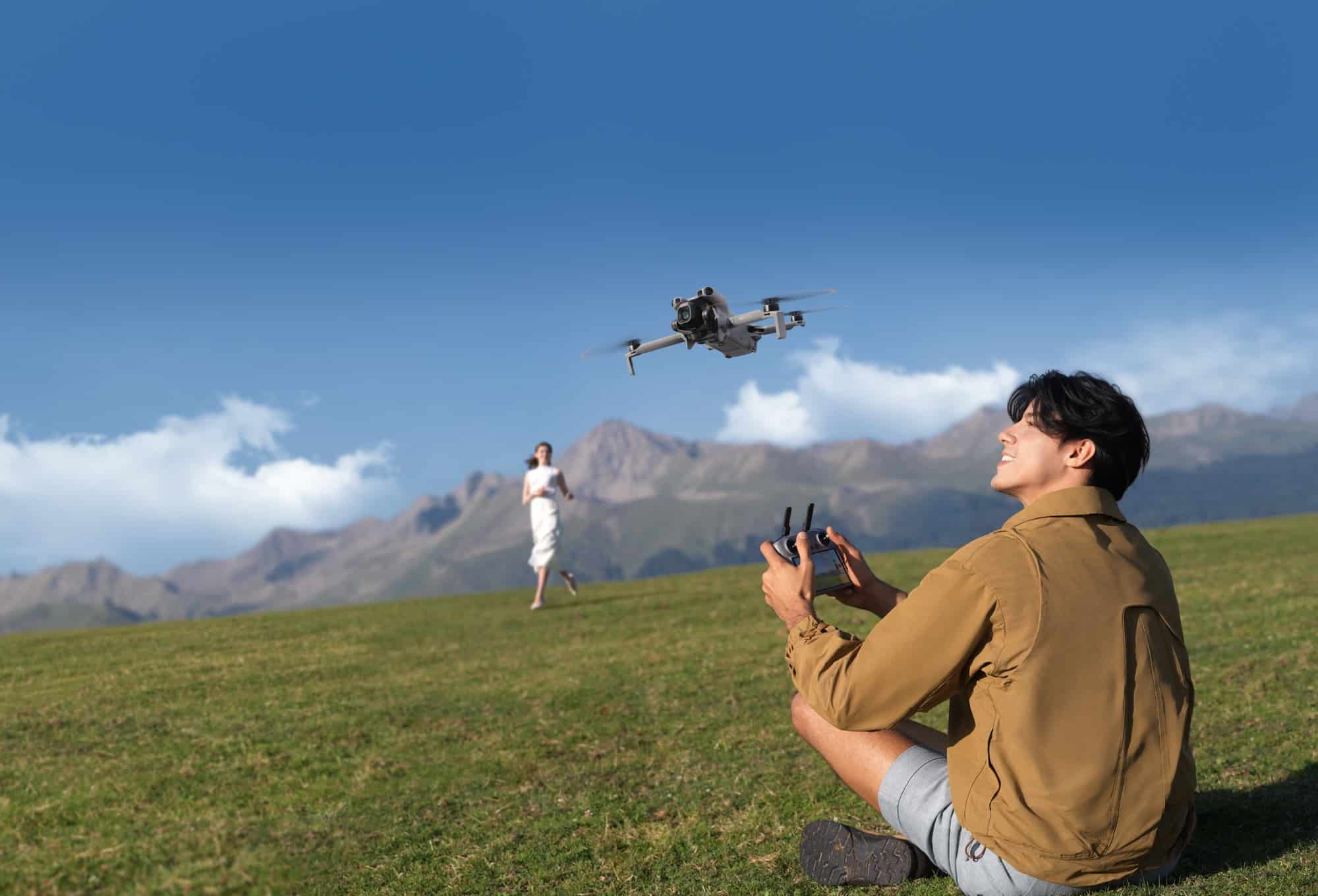 A man sits on a grassy slope flying toward a woman under towering clouds.