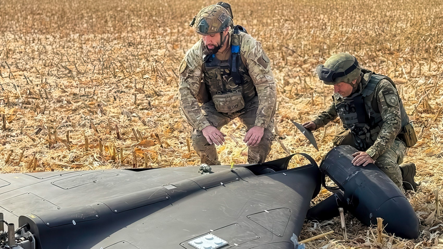Soldiers examining a downed delta-wing attack drone in a harvested field.