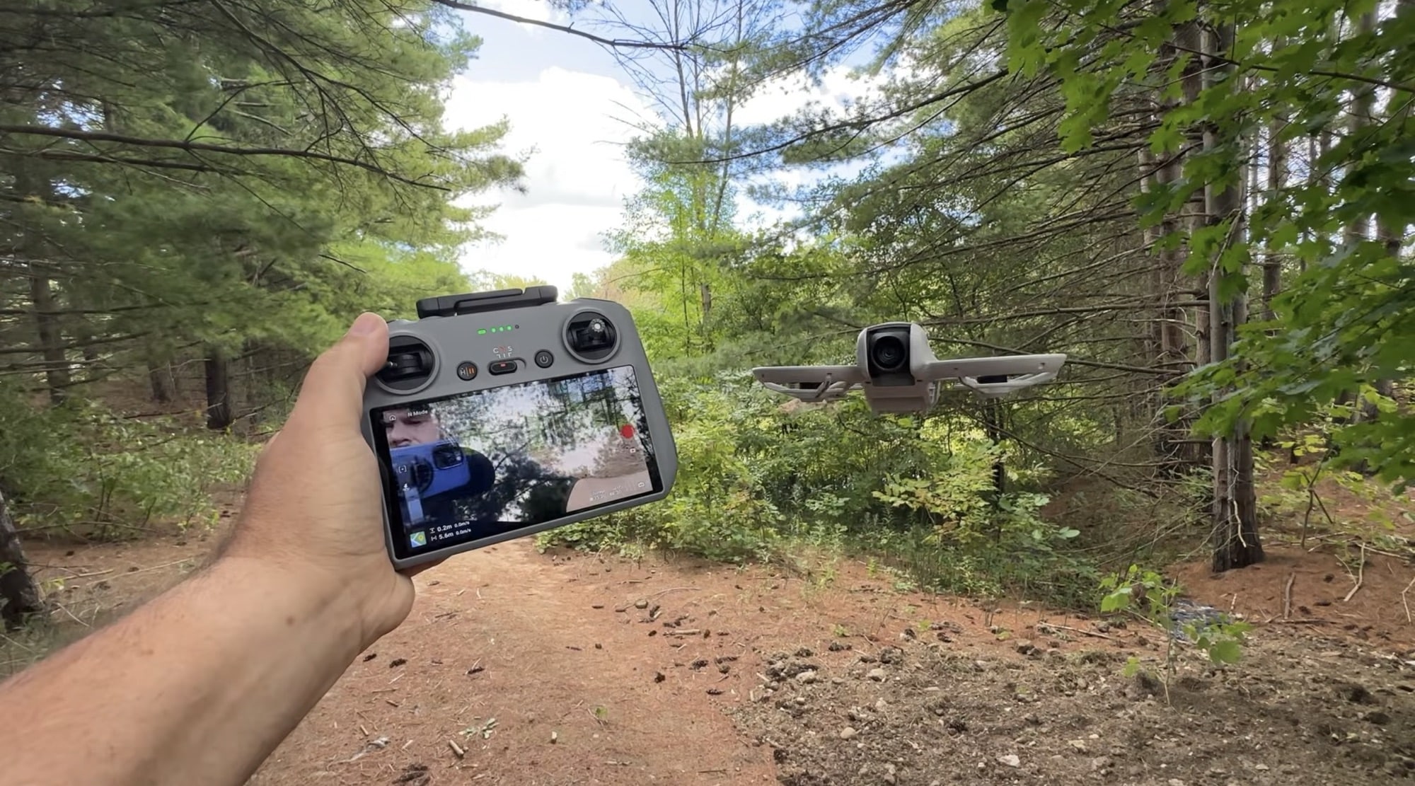 A pilot holds a screen controller as the Neo hovers above a wooded path.