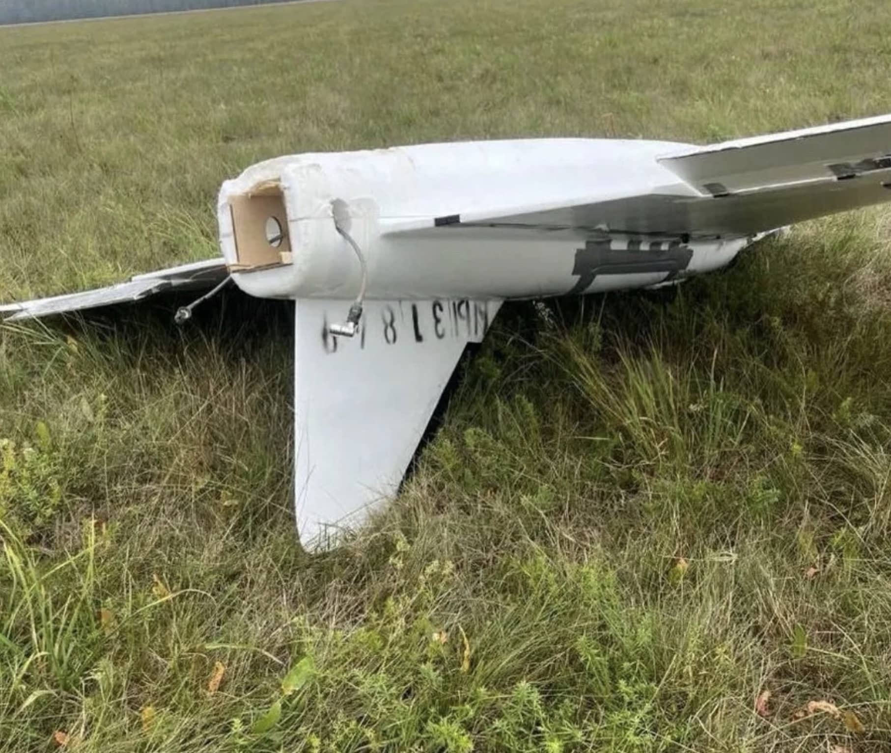 White foam fixed-wing drone with hand-painted serial numbers lying intact in tall grass.