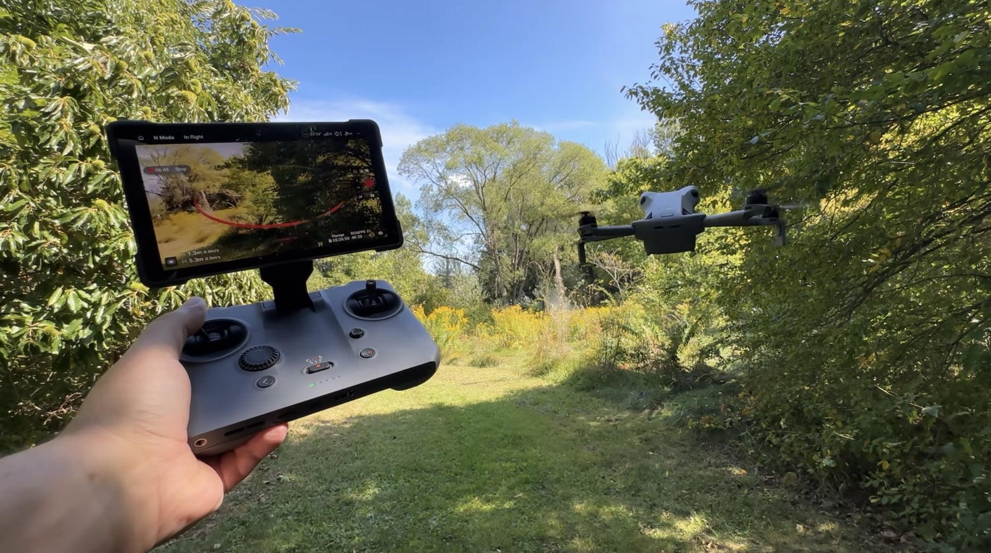 DJI Mini 5 Pro hovering above a grassy clearing while the pilot holds the controller