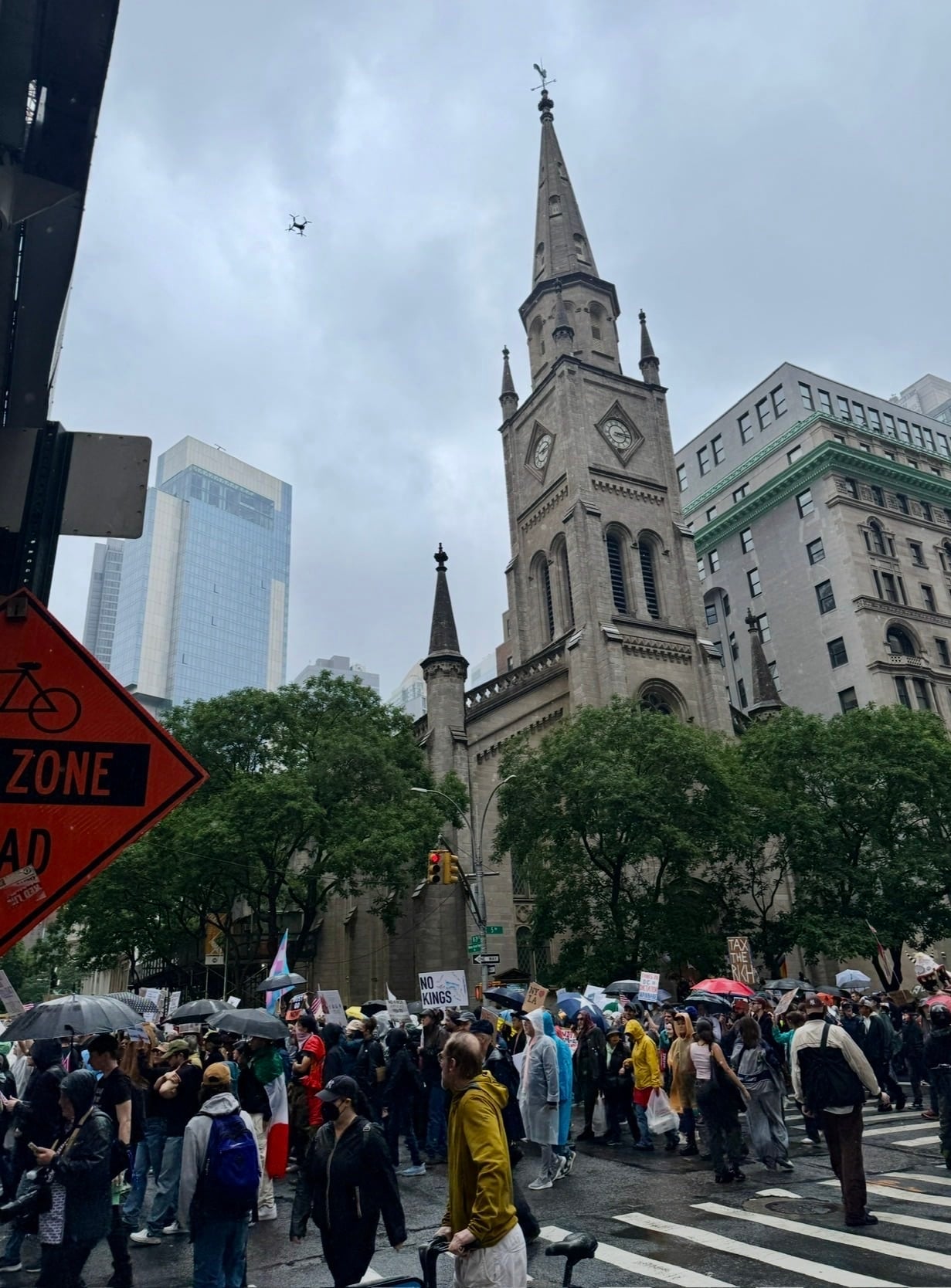 A drone hovers above a rainy protest crowd near a stone church spire.