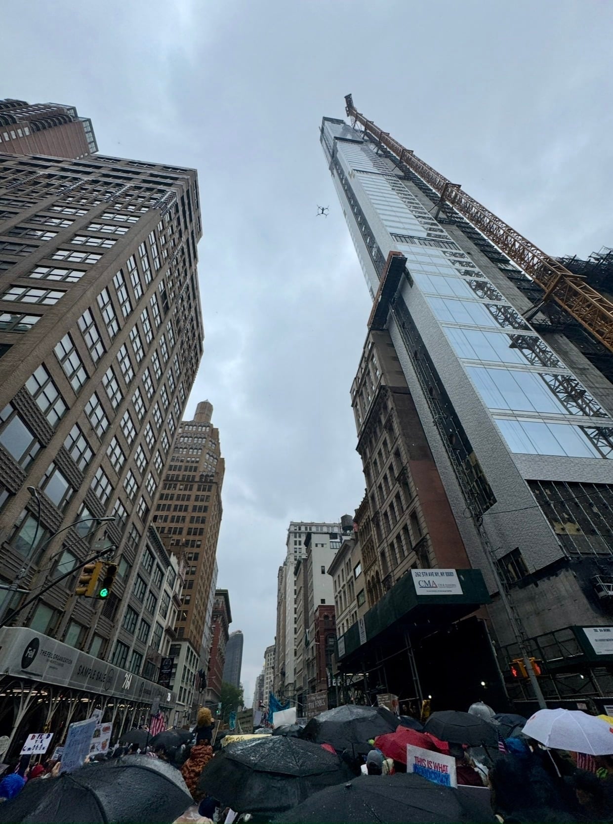 Looking up a narrow city canyon at a distant drone above umbrella-covered marchers.