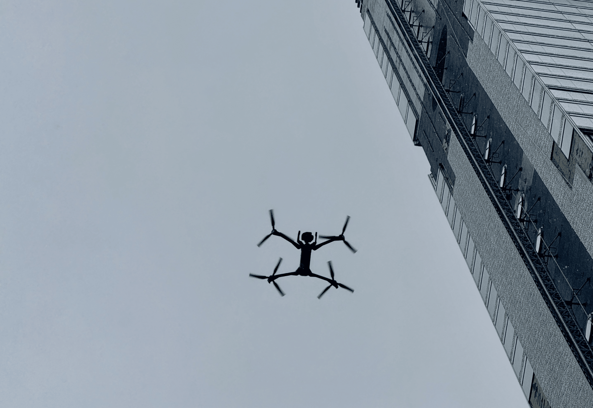 An X-shaped quadcopter hovers between office towers under a flat grey sky.