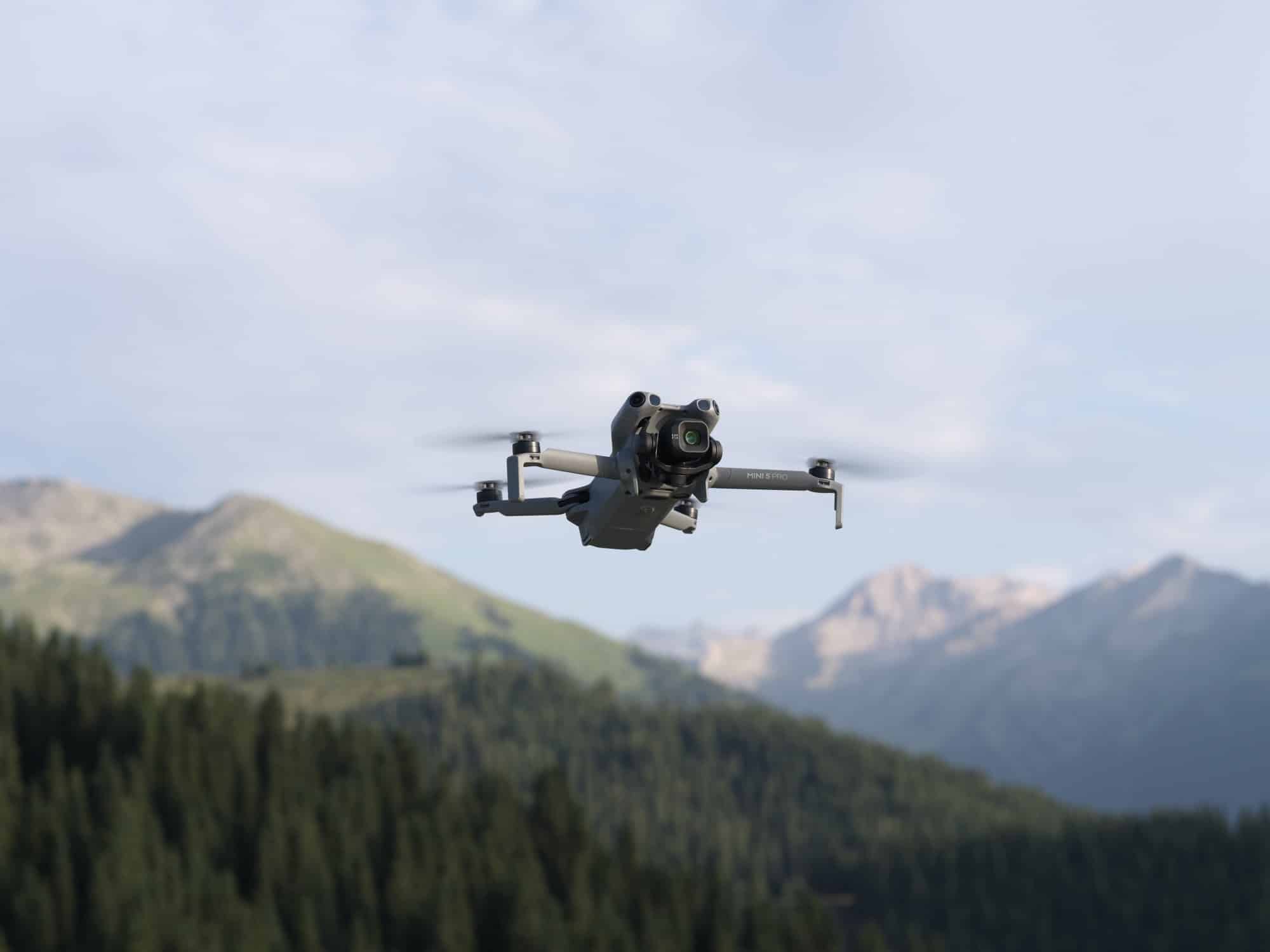 The drone hovers above a forested valley with bare mountain ridges rising beyond.