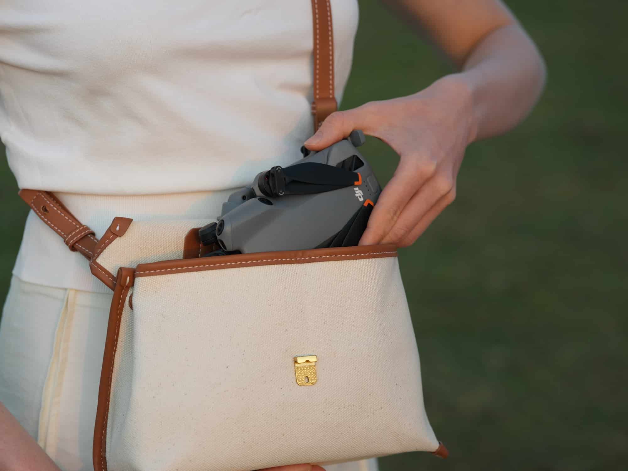 A woman lowers the folded drone into a cream canvas crossbody bag.