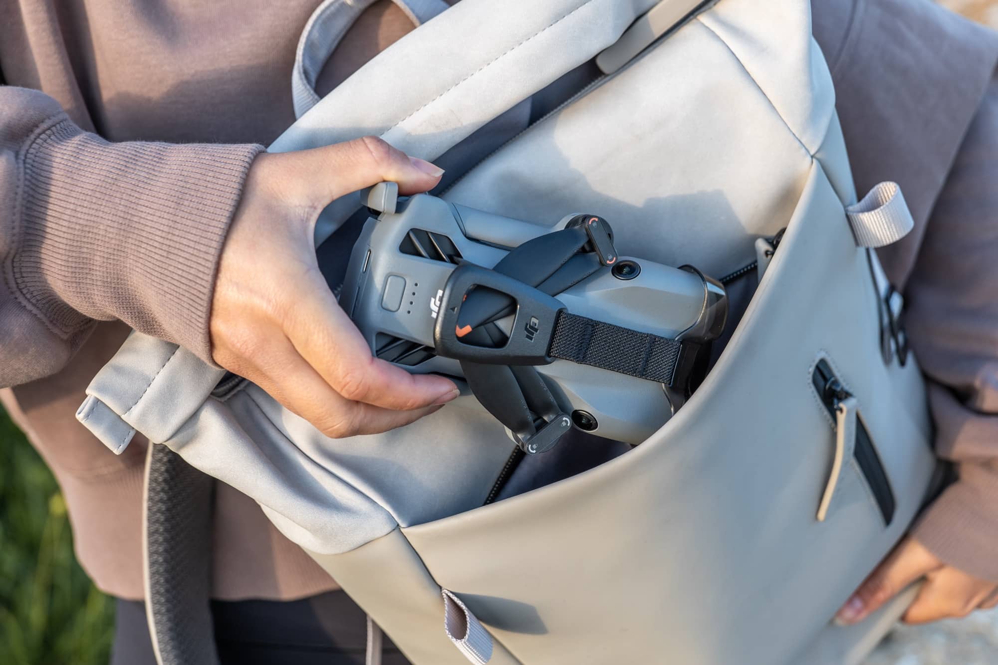 Hands tuck the folded drone into the side pocket of a pale shoulder bag.
