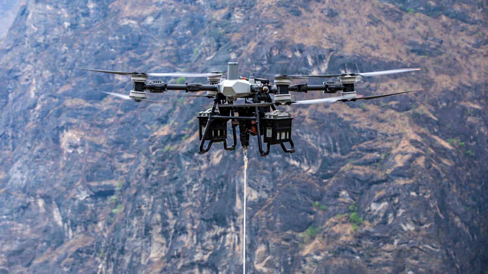 DJI FlyCart 100 heavy-lift drone hovering above rocky terrain with winch line deployed.