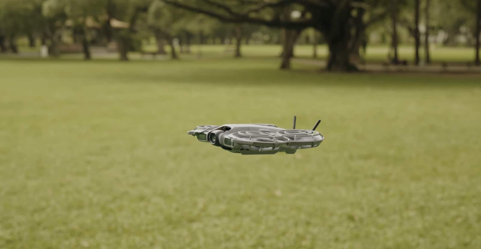 The drone skims low over a mown park lawn ringed by shade trees.