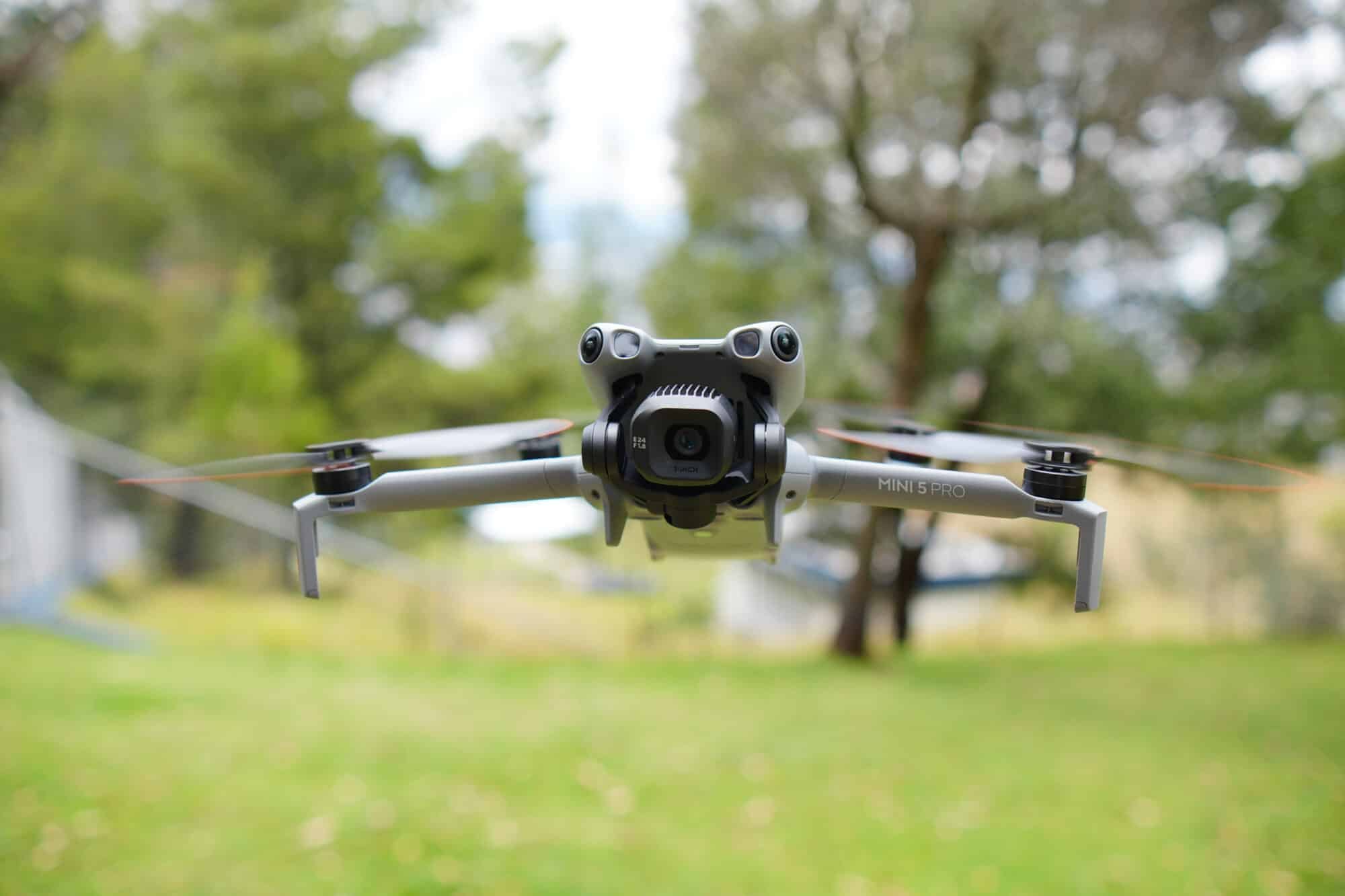 DJI Mini 5 Pro hovering low above a sunlit lawn with trees behind.