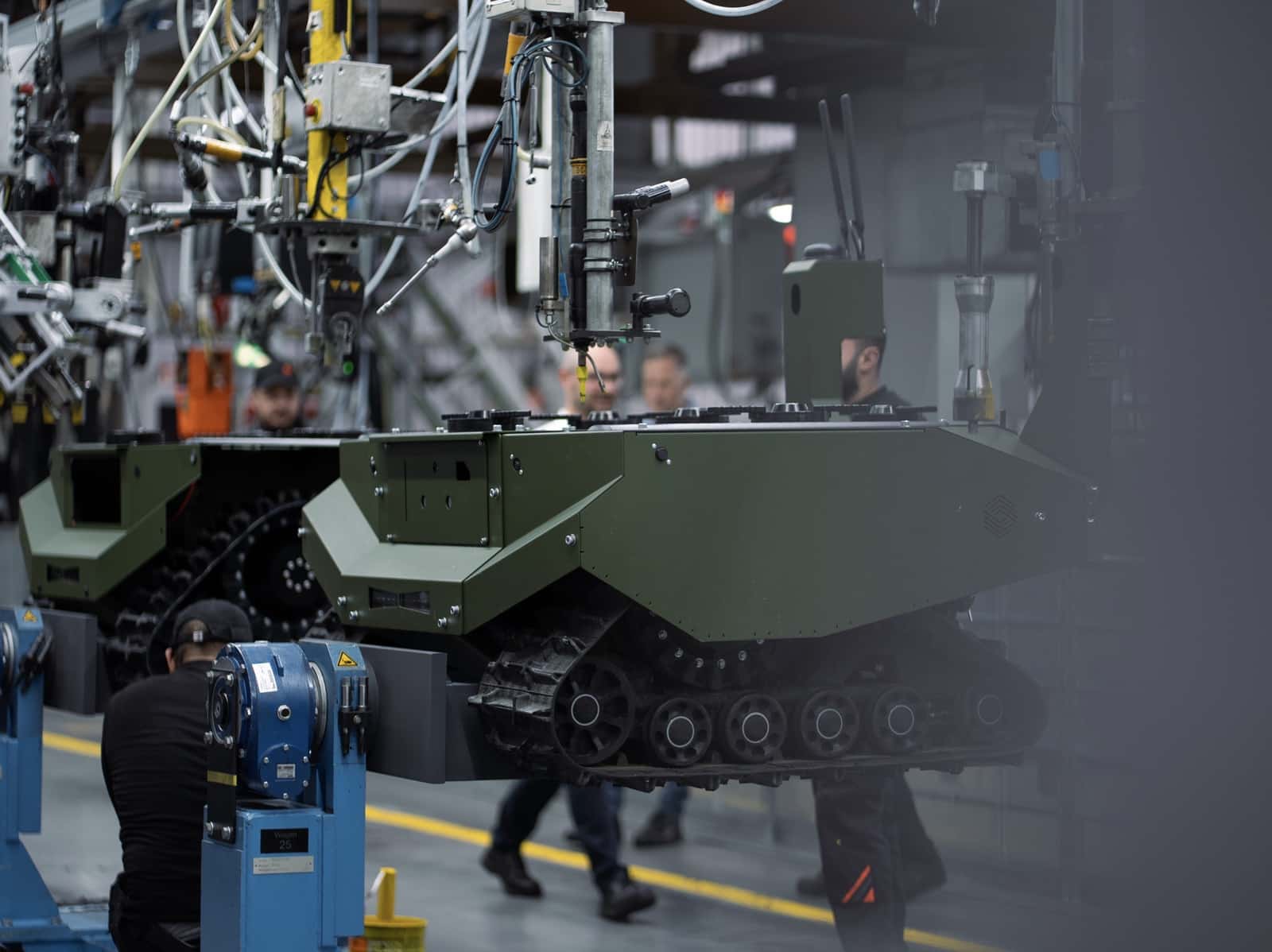 Green tracked unmanned ground vehicle moving along an assembly line with workers.