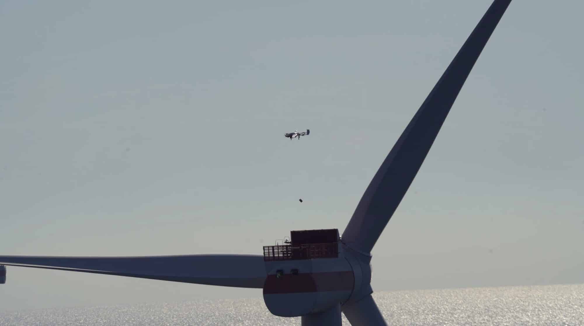 Distant drone flying near an offshore wind turbine blade against pale sky.