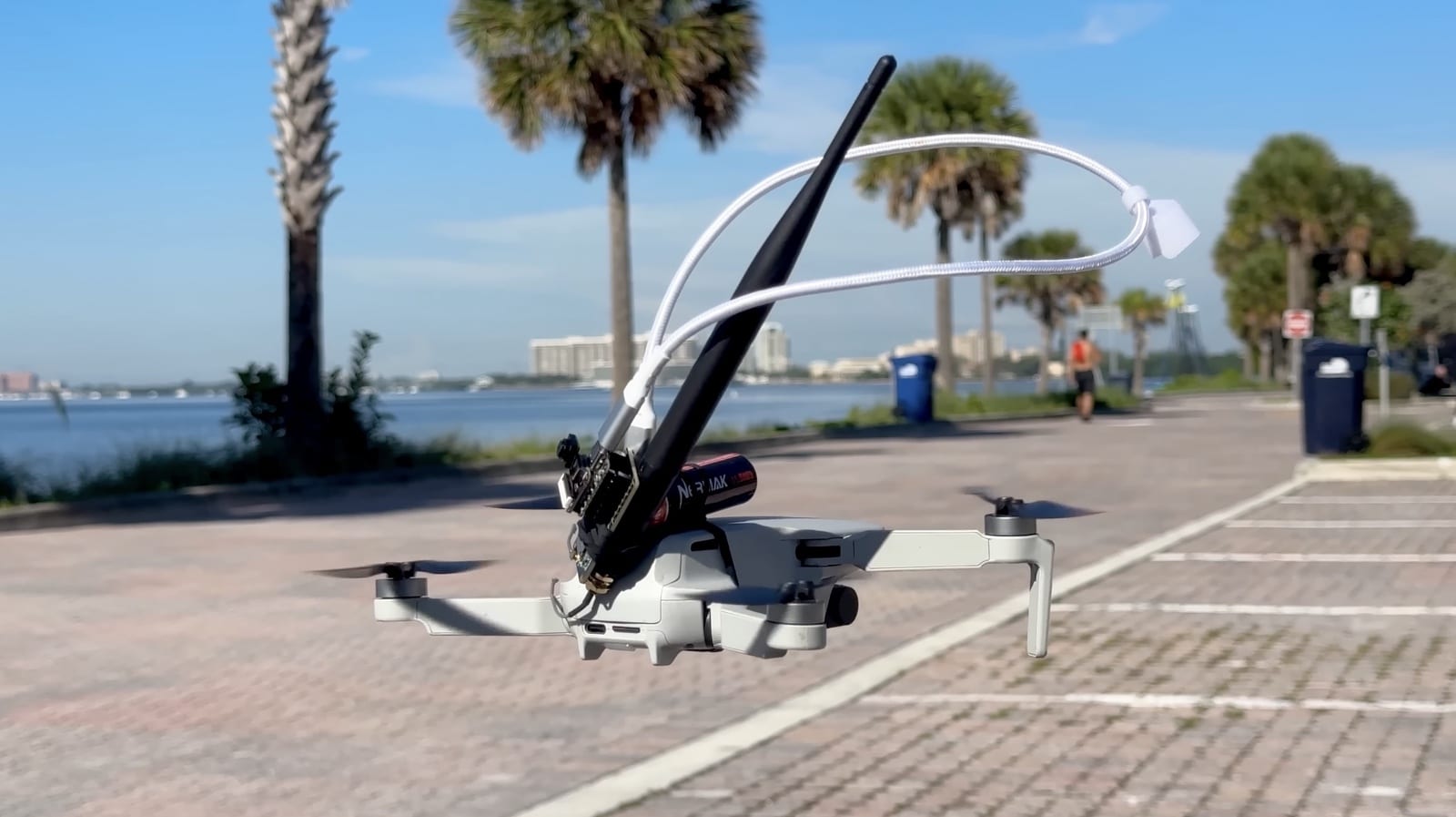 A small quadcopter carries a strapped-on antenna payload on a waterfront promenade.