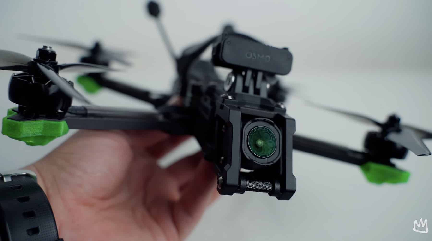 A hand holds an FPV freestyle quadcopter with green propellers up to the camera.