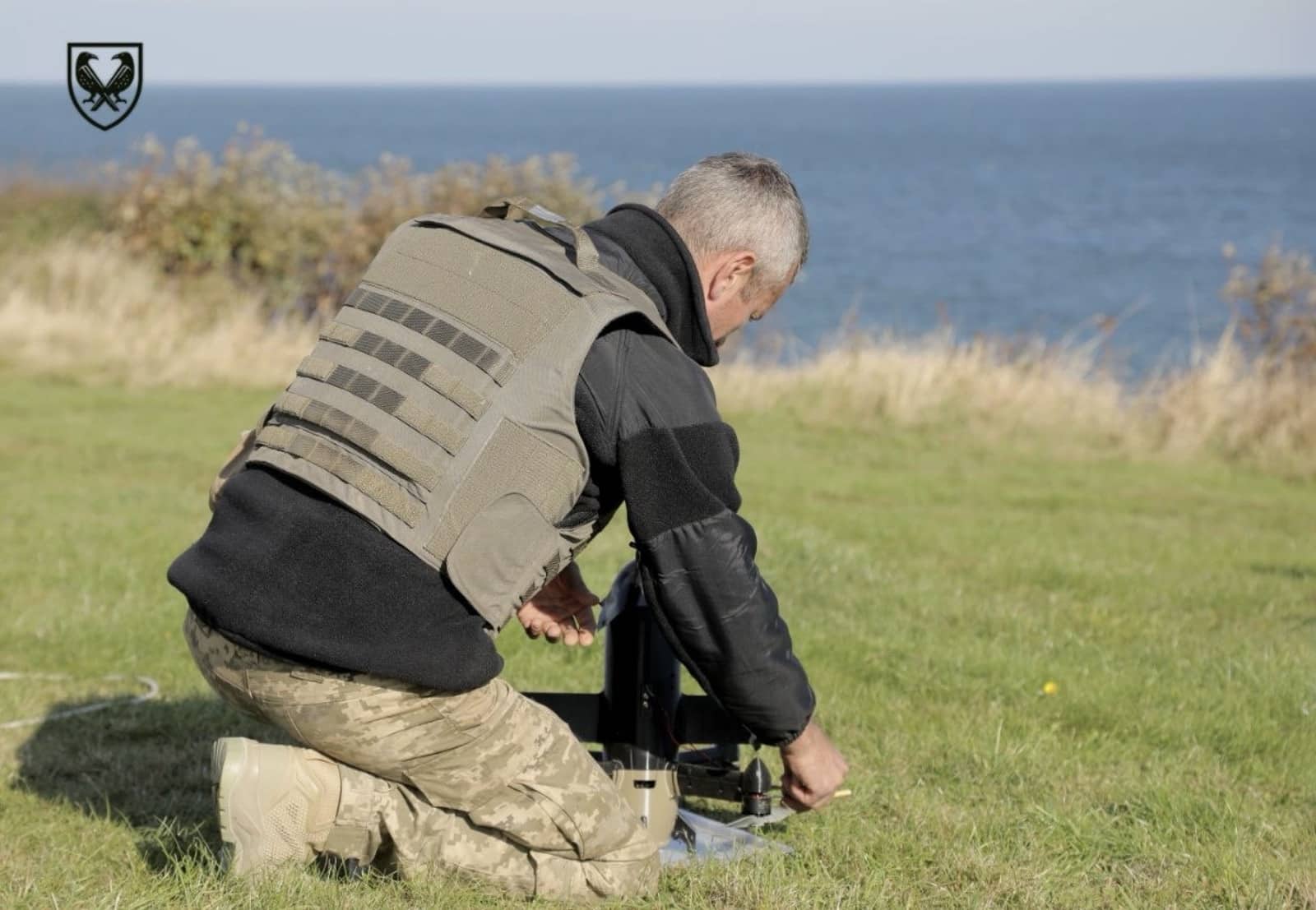 A man in body armor kneels on coastal grass adjusting a drone launch rail.