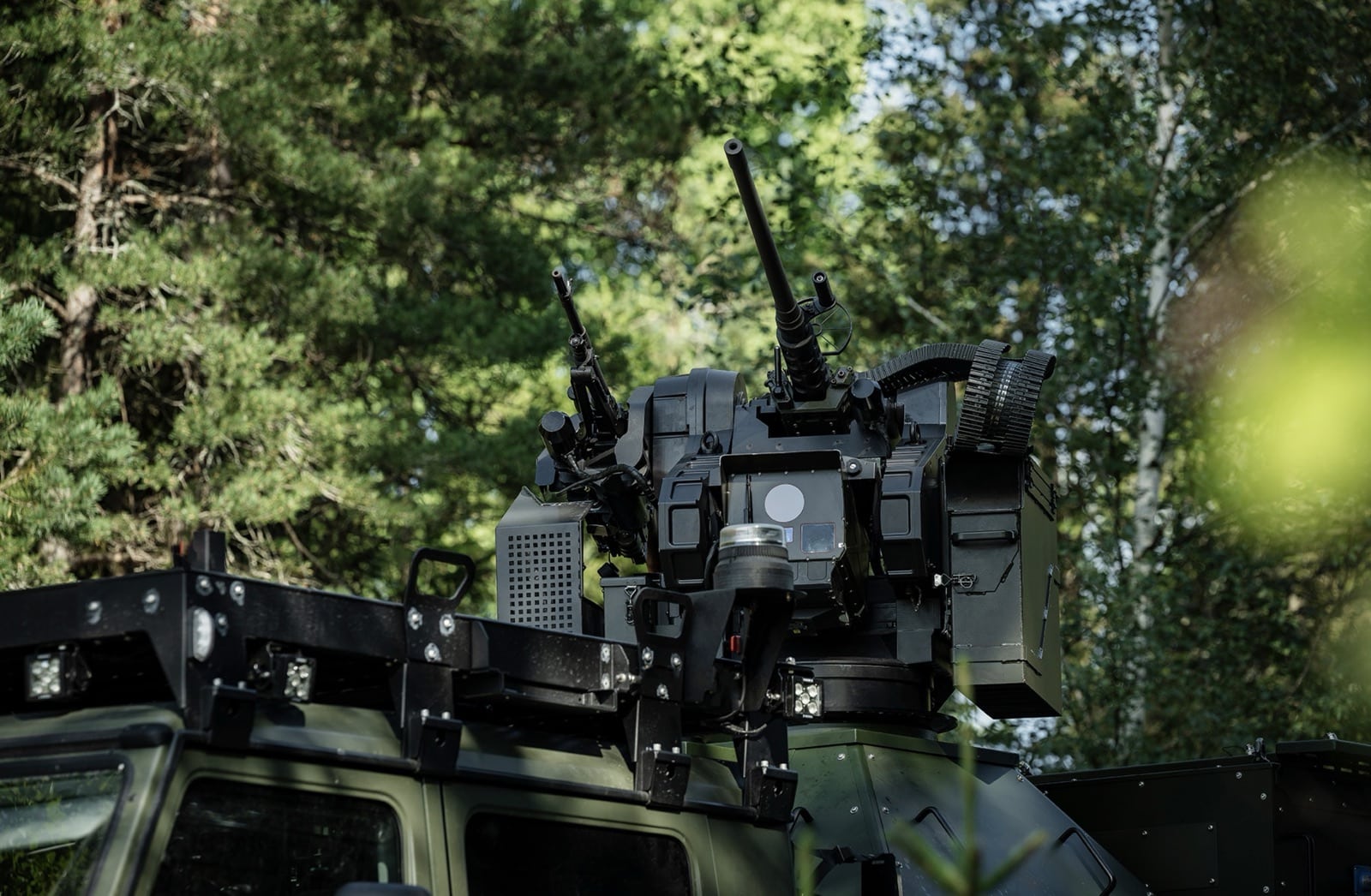 Remote weapon station mounted on an armored vehicle roof among trees.