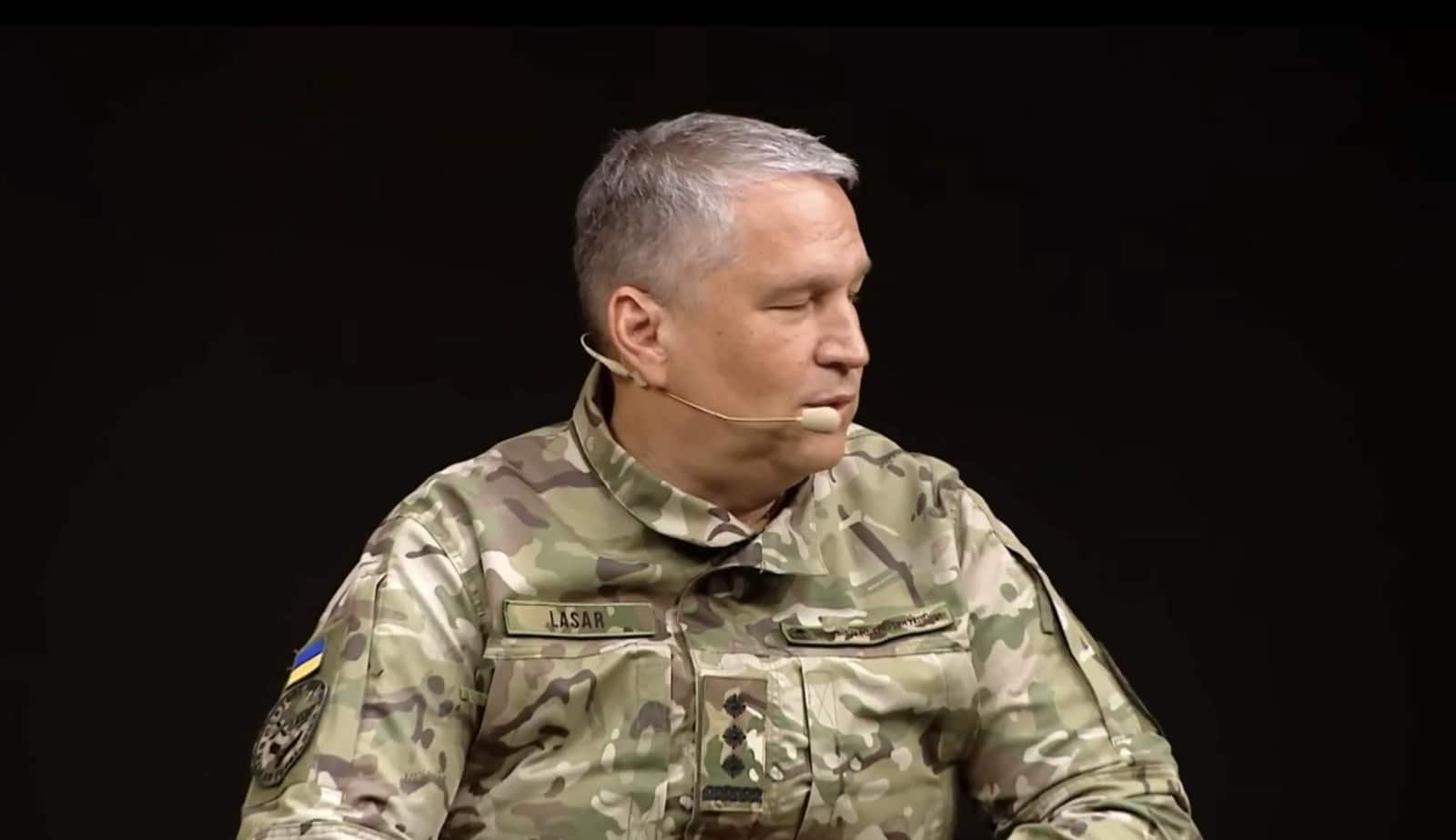 A man in Ukrainian military uniform with a headset speaking against a dark background.
