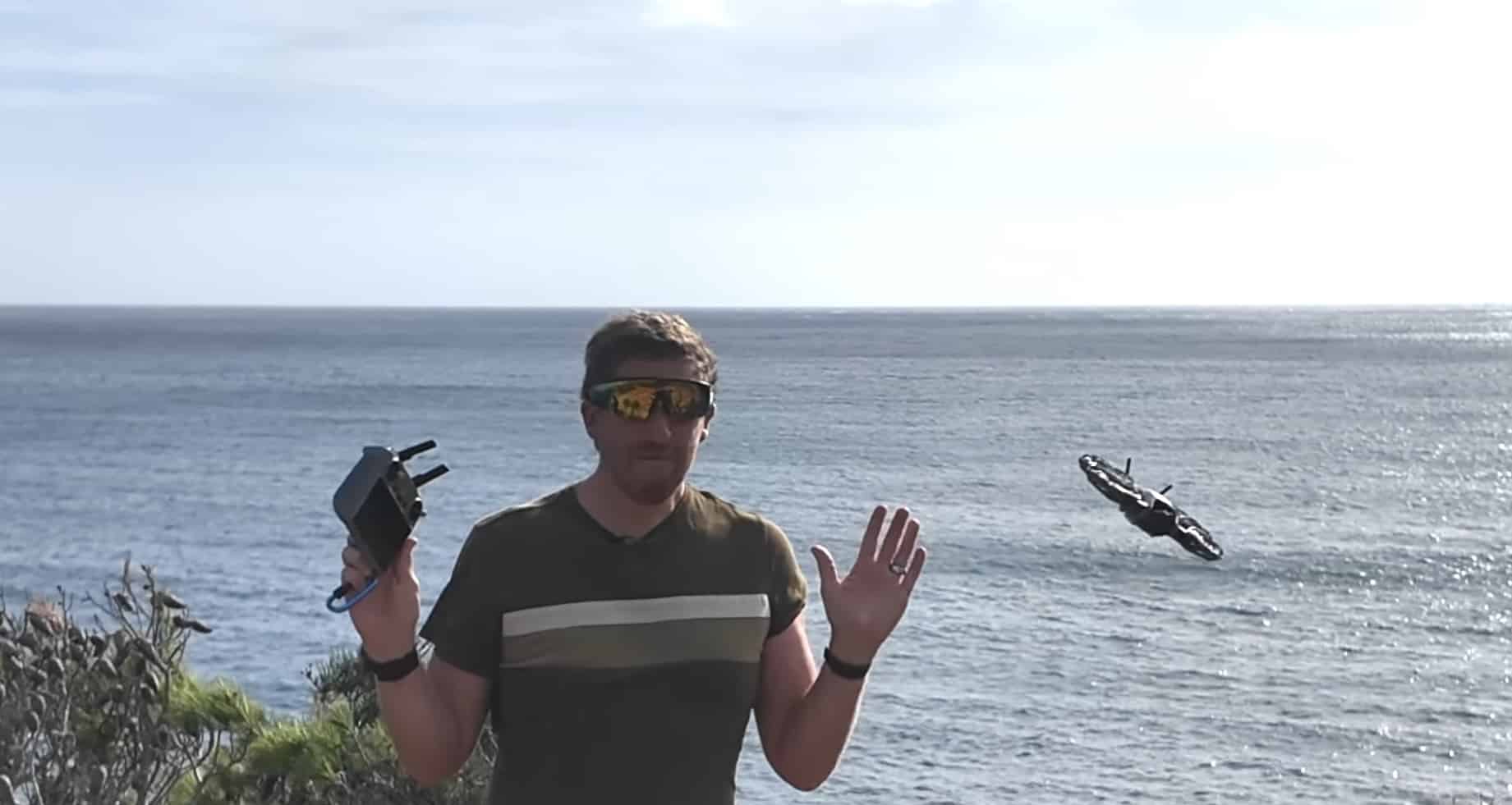 Pilot with controller on a windy clifftop as a drone hovers over the sea.