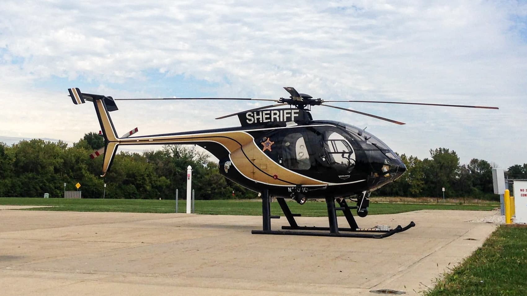 Sheriff department helicopter parked on a paved ramp beside a treeline.