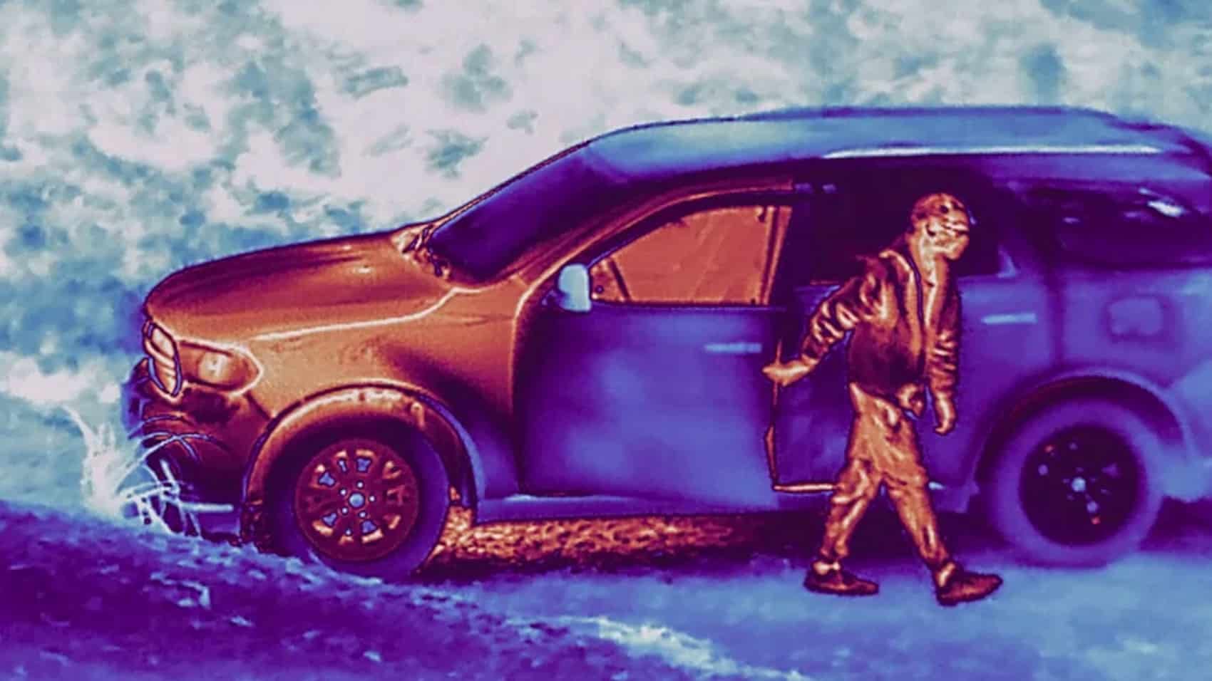 Thermal image showing a person walking past a parked SUV in false color.